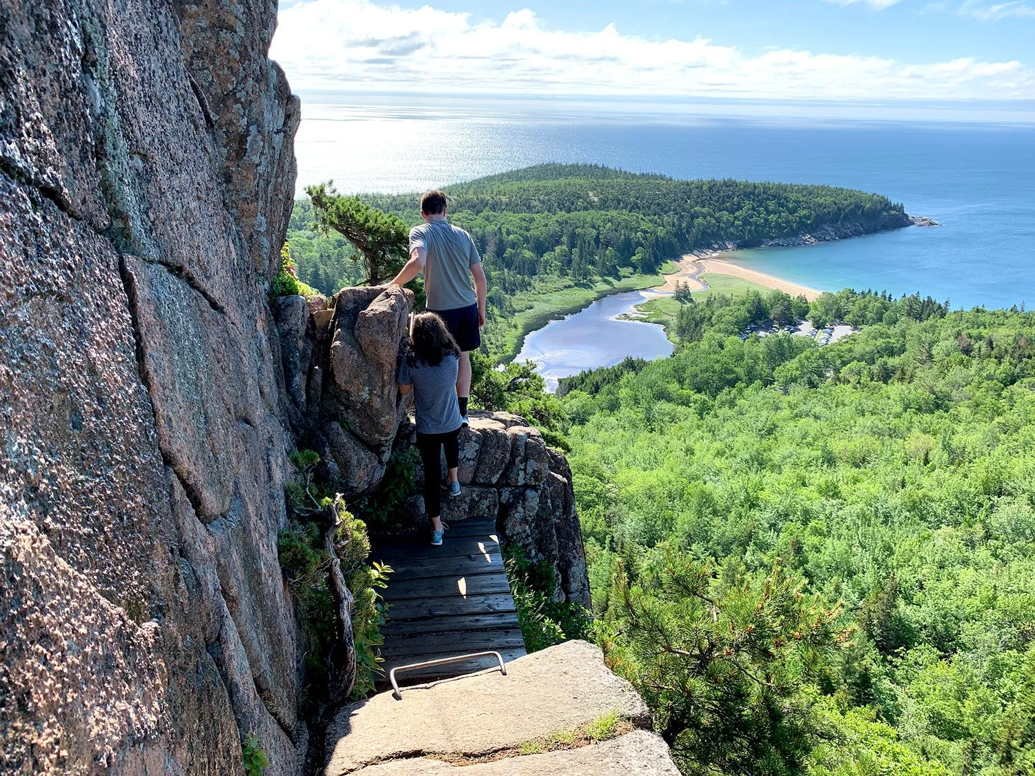 Two people hiking on a narrow wooden trail along a rocky cliff, overlooking a lush green forested landscape, a lake, and a beach by the ocean.