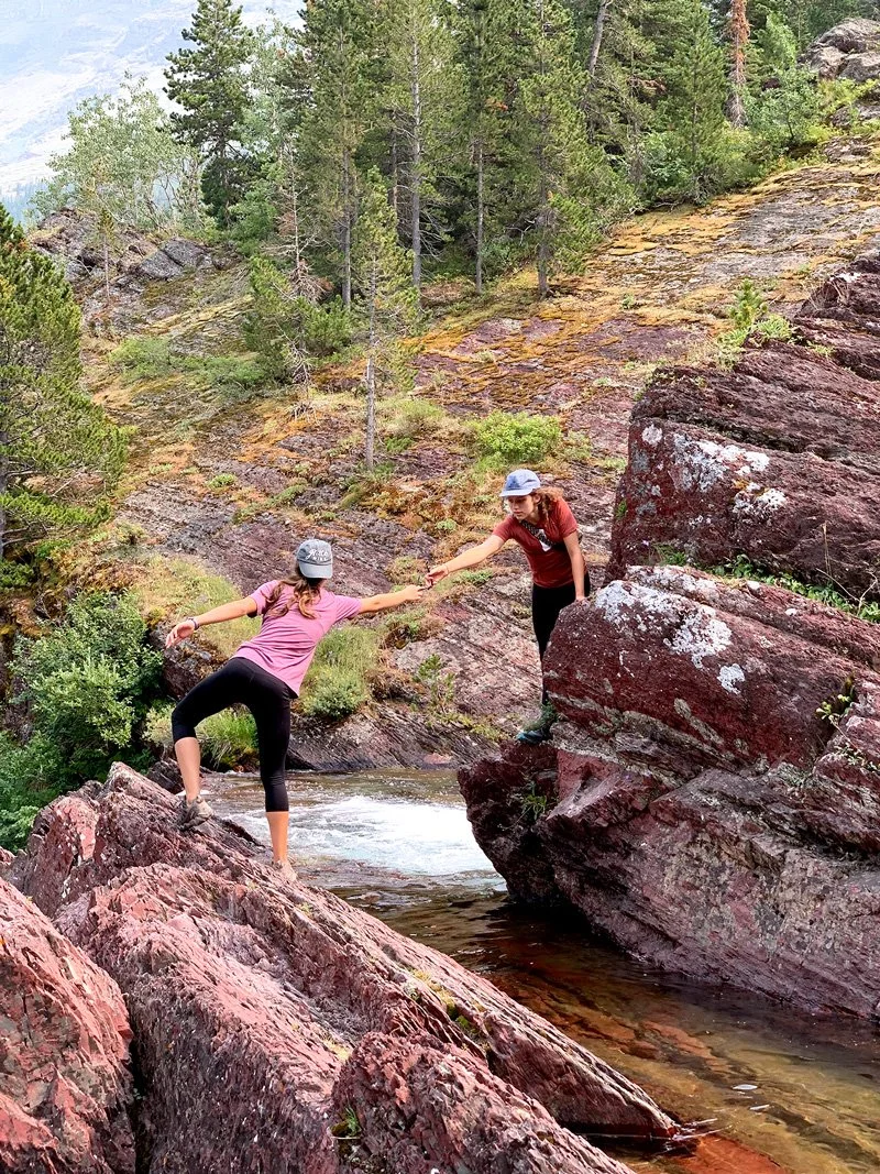 Two women, one in a pink shirt and the other in a red shirt, helping each other across rocks by a shallow river in a forested area.