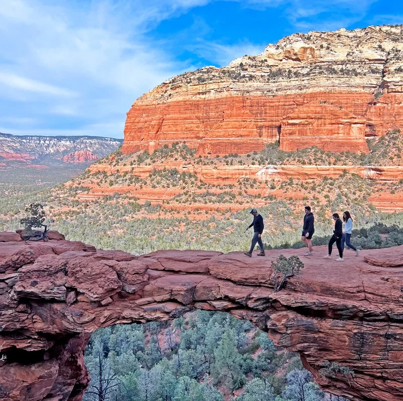 Four people hiking on a red rock trail in a canyon, with a large red and orange layered cliff in the background under a partly cloudy sky.