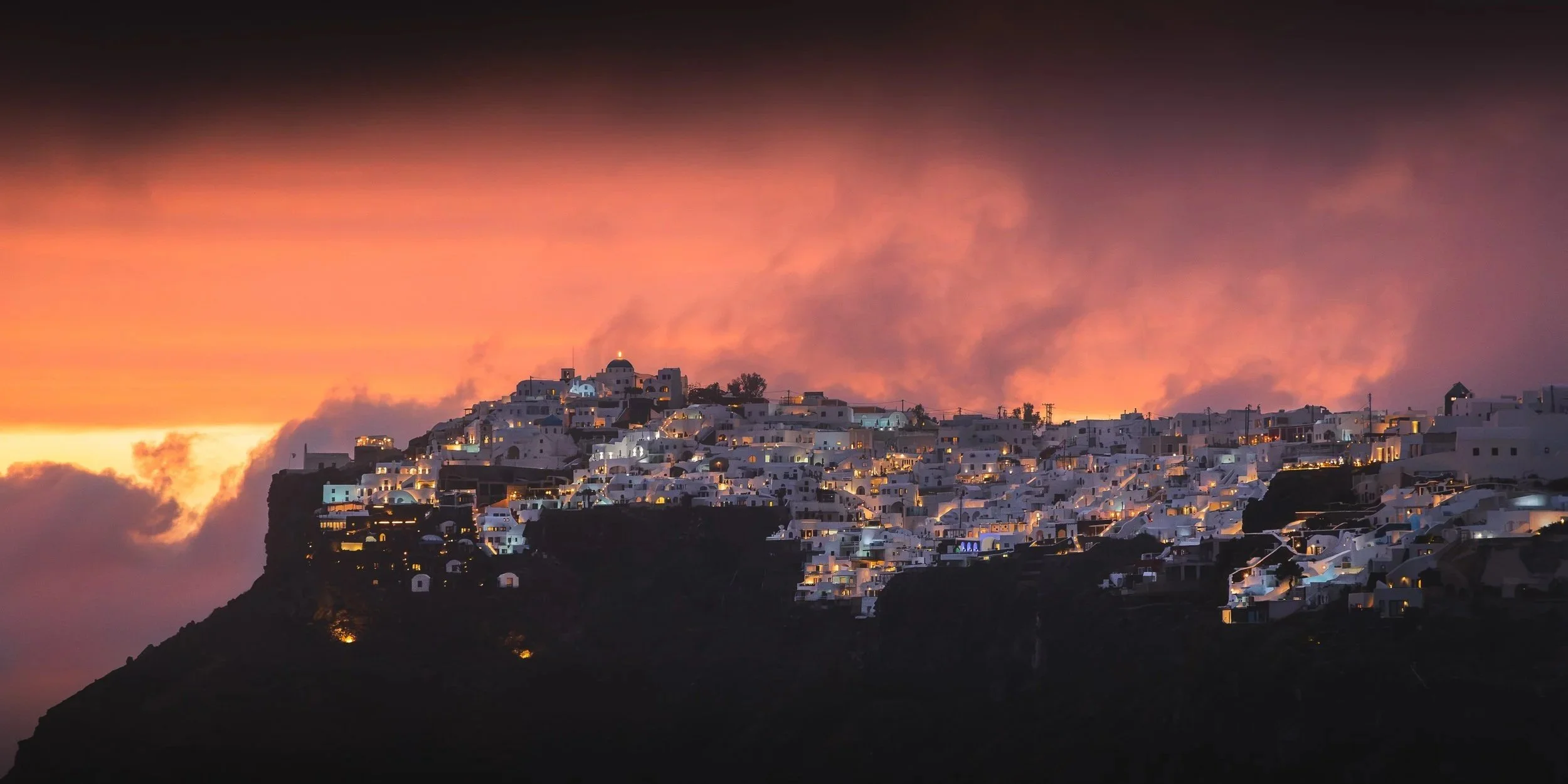 Whitewashed buildings of Santorini, illuminated beneath a dramatic pink and orange sunset sky over the Aegean Sea.