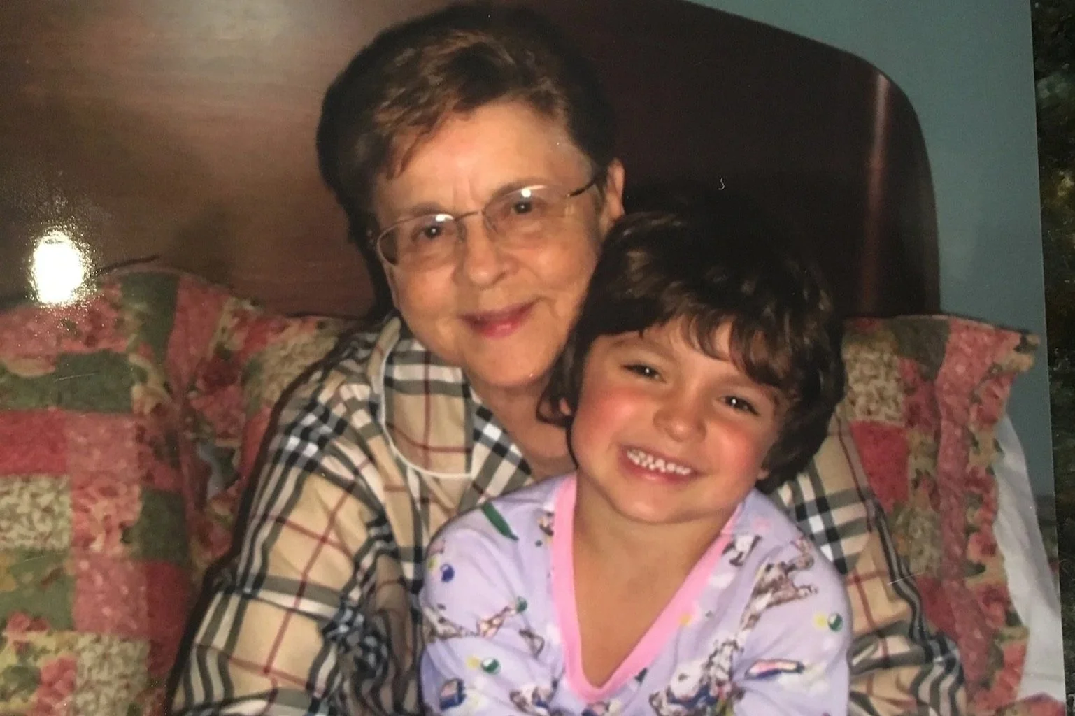 A photo of an elderly woman and a young boy smiling together, sitting on a floral couch with pillows behind them.