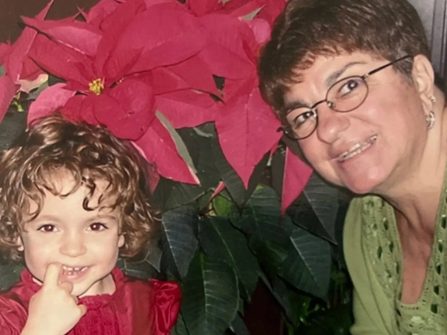 A young girl with curly hair smiling and touching her chin, and an older woman with short hair, glasses, and earrings, smiling, standing next to a poinsettia plant with red leaves.