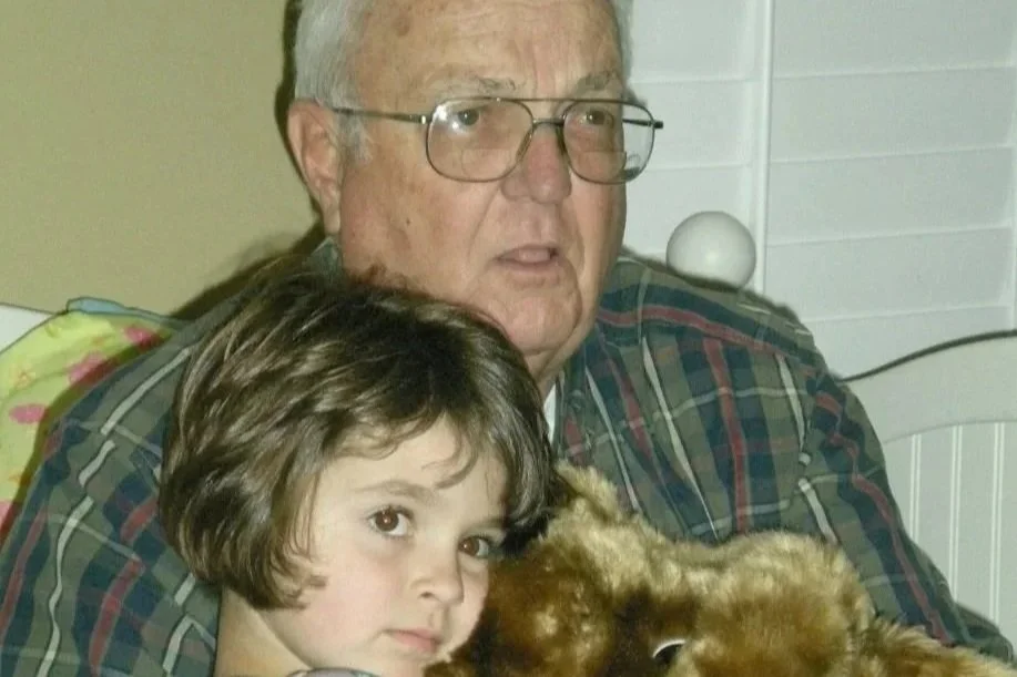 A young girl with brown hair and an older man with glasses sitting together, with a brown animal, possibly a dog or a stuffed animal, partially visible in the foreground.