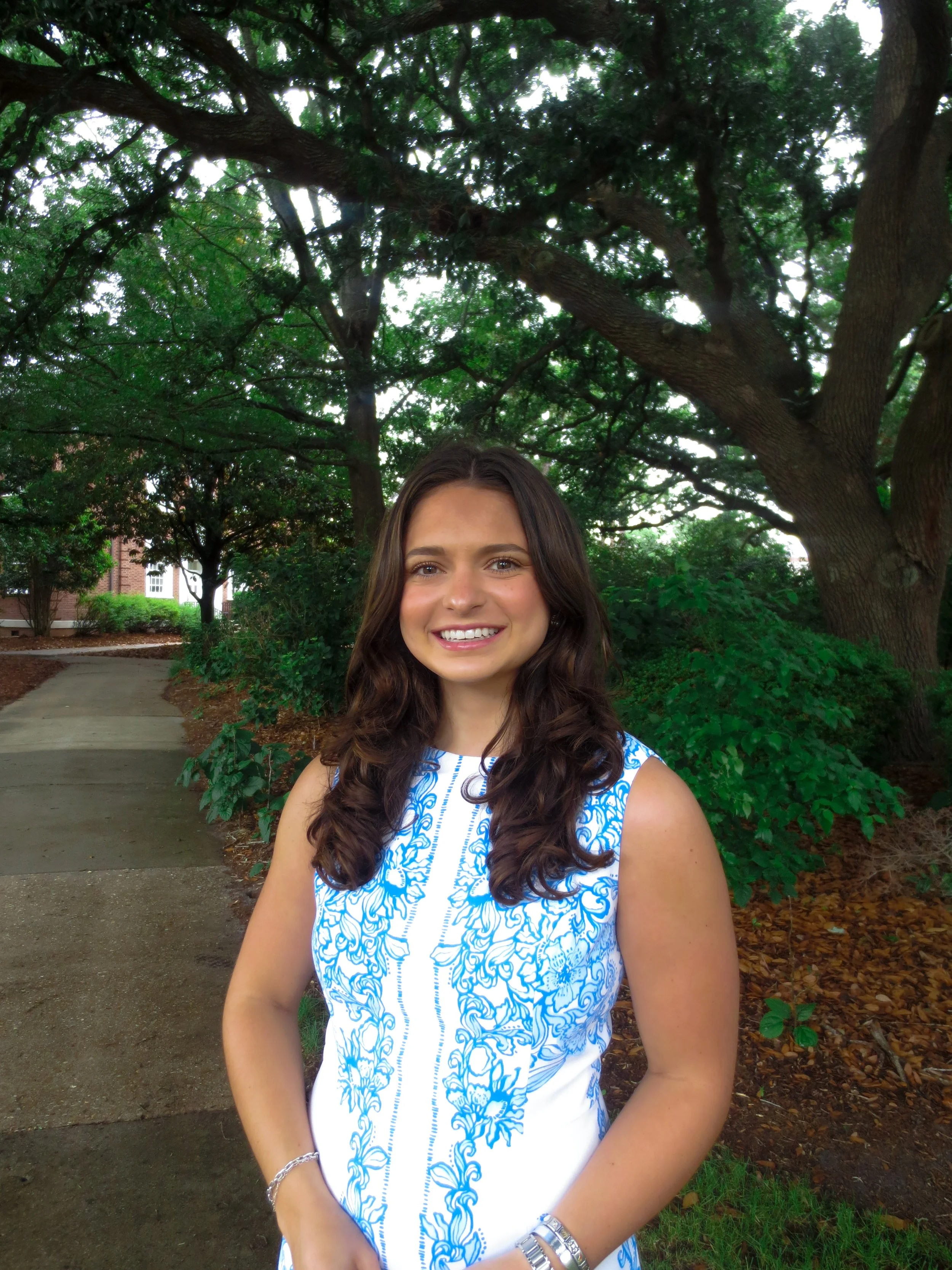 A young woman with long brown hair and a bright smile standing outdoors in a park, wearing a white and blue patterned sleeveless dress, with trees and greenery in the background.