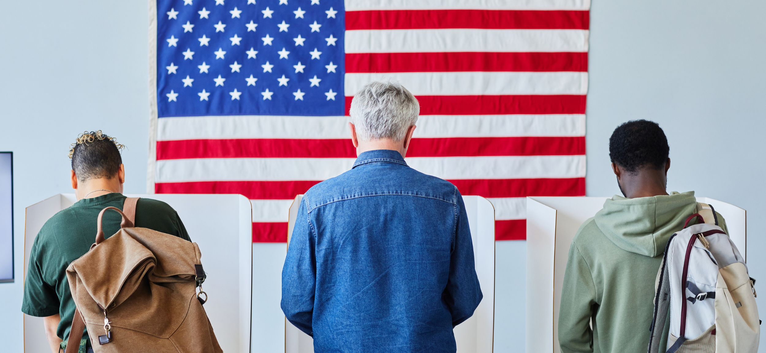 Three people standing in front of an American flag, with their backs facing the camera, possibly voting or at a civic event.