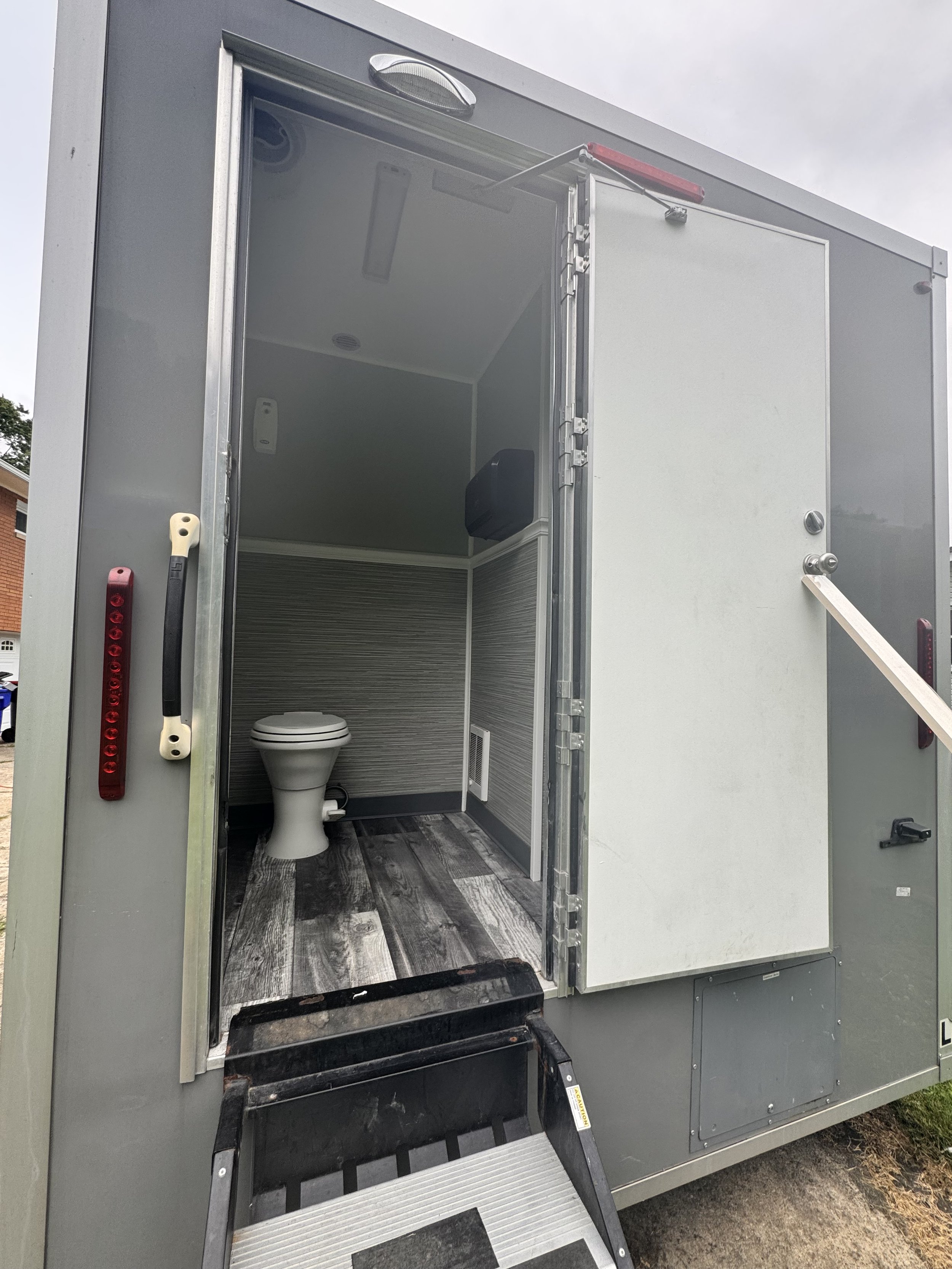 The image shows a restroom inside a mobile or portable unit, featuring a white toilet, gray and white interior walls, and a wood-patterned floor. There is a step ladder outside the entrance.