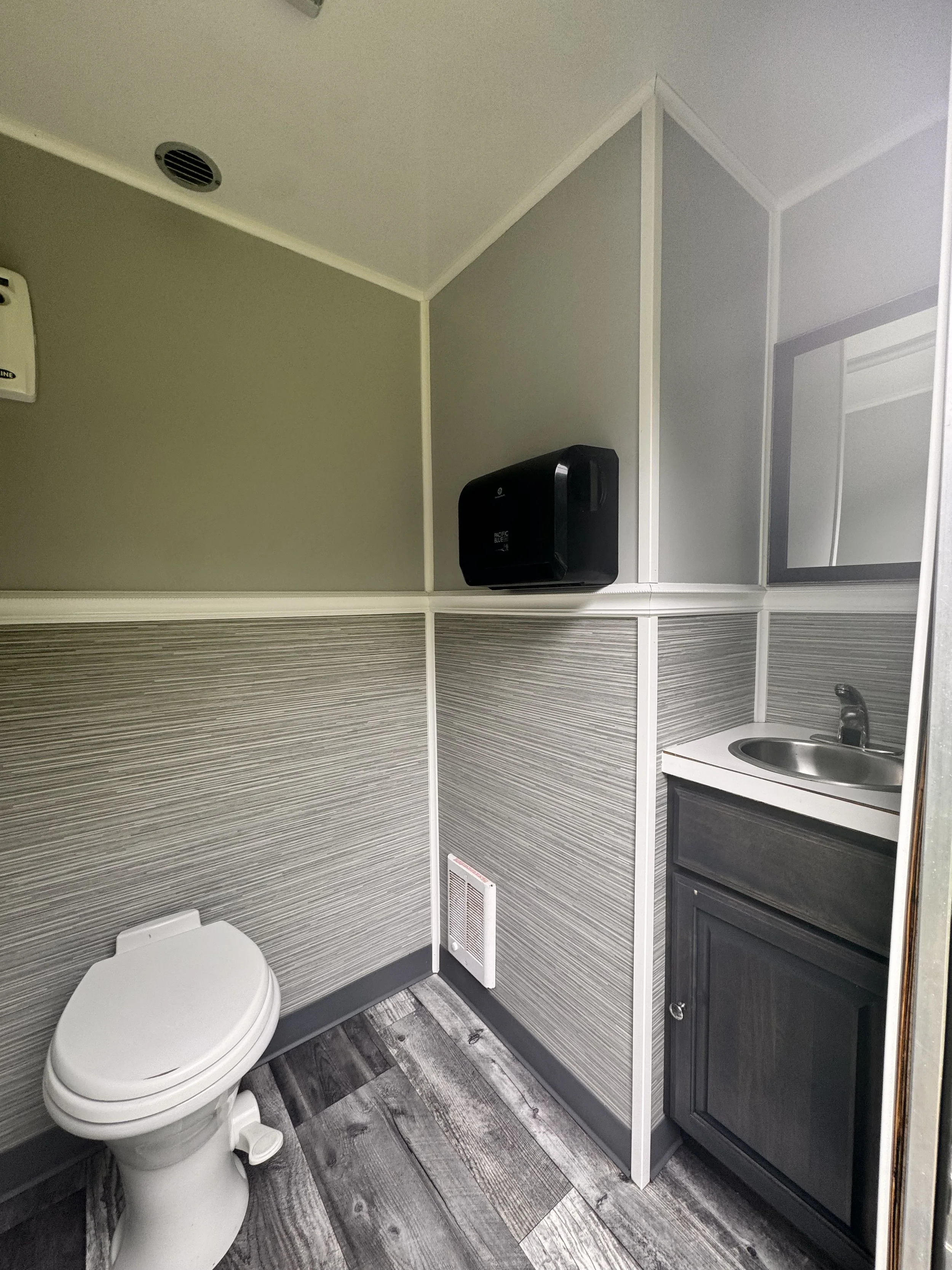 A small public restroom with a white toilet, gray panels, a wall-mounted black paper towel dispenser, a mirror, and a small sink with dark cabinetry. The floor has gray textured tiles.