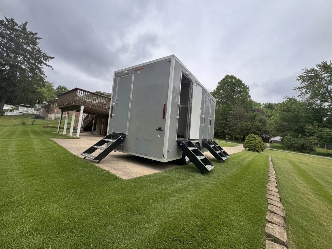 A large gray mobile trailer with stairs leading to doors, situated on a grassy lawn in a suburban neighborhood with trees and houses in the background.