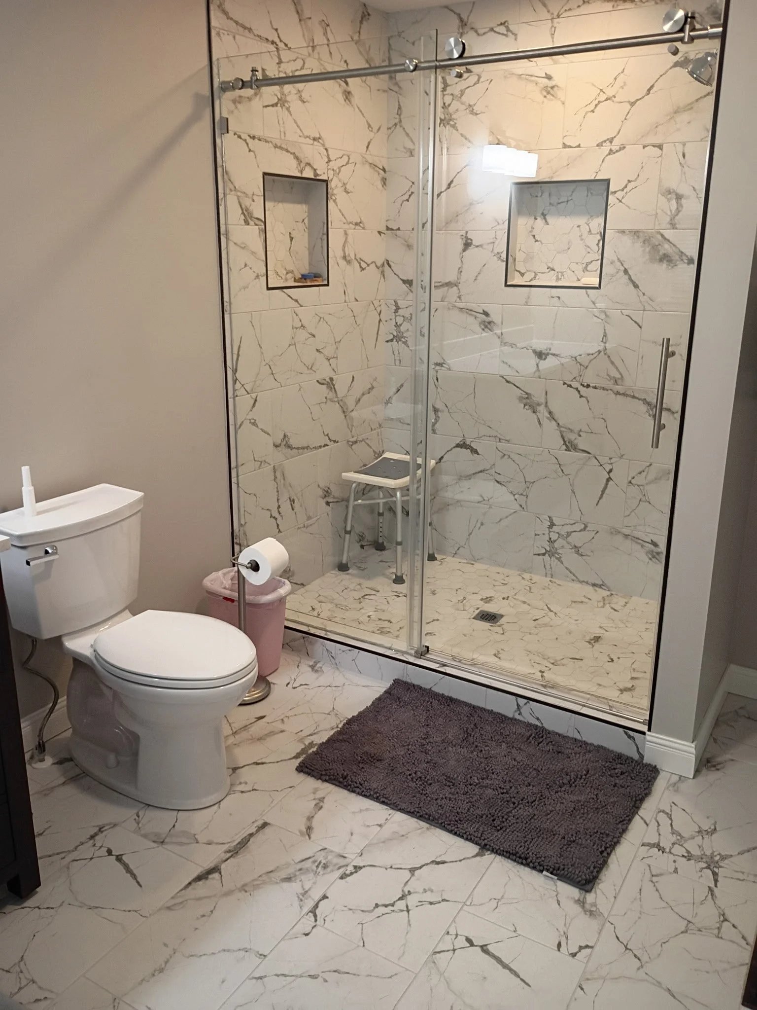 A bathroom with marble tile flooring and a marble tiled shower enclosure with a glass door, two recessed shelves, and a shower chair. A toilet, a toilet paper holder, a wastebasket, and a brown bath mat are also visible.