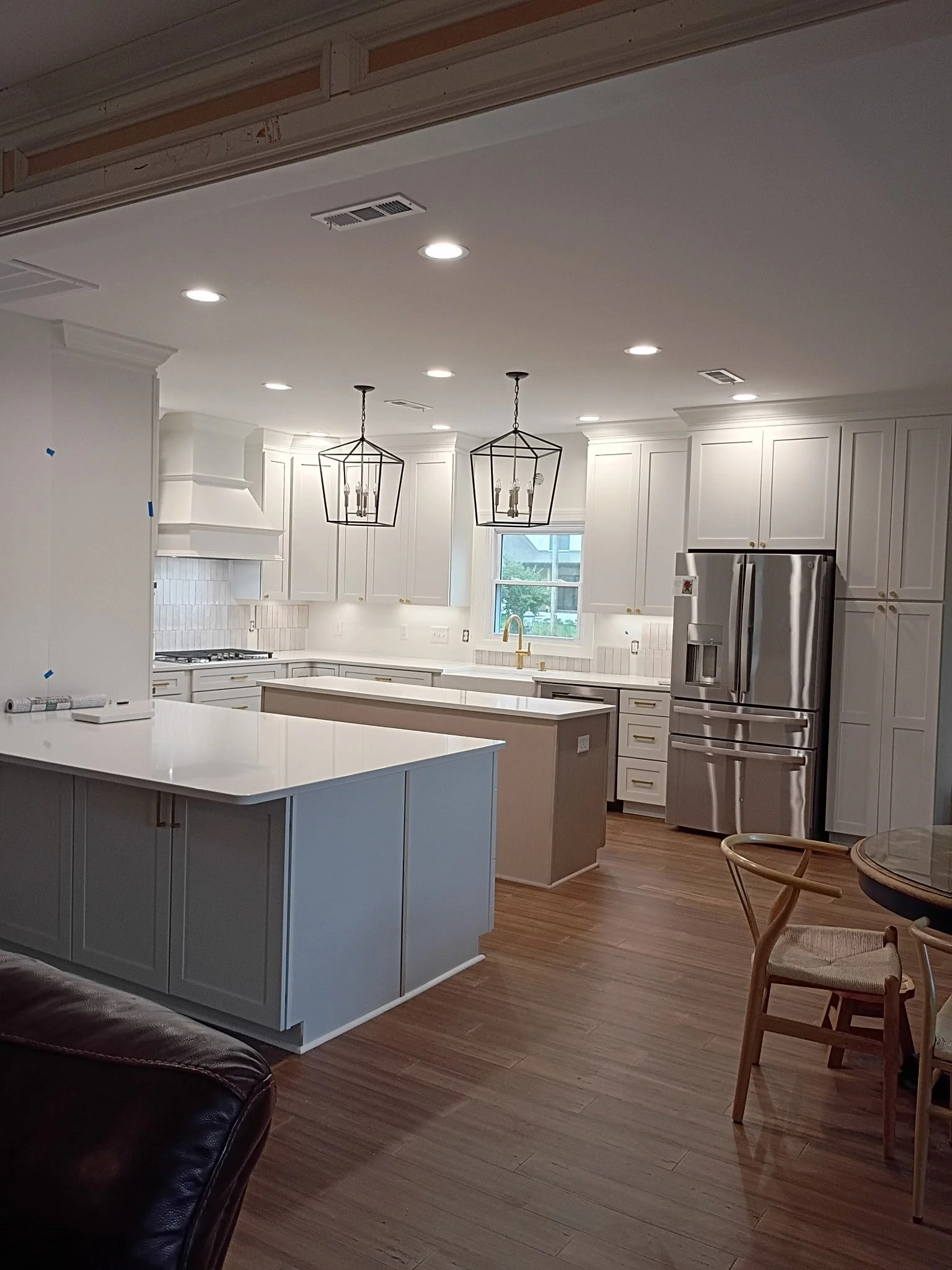 Interior view of a kitchen under renovation with white cabinets, a island, pendant lights, and stainless steel refrigerator, seen from a living room with a leather couch and a wooden chair.