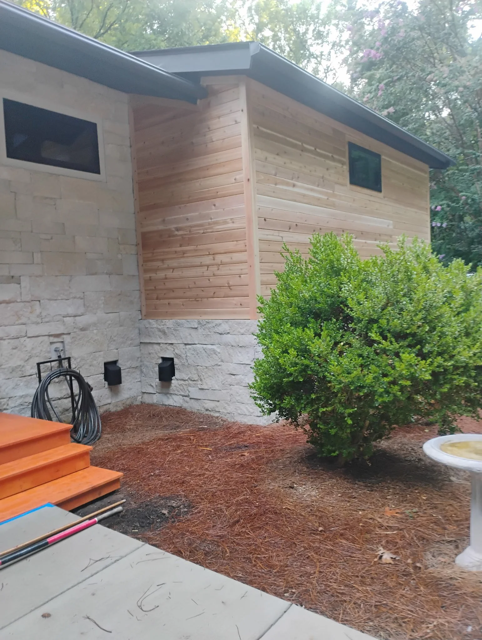The image shows the backyard of a house with a staircase on the left, a hedge on the right, and a house wall with two small windows. There are some garden tools and hoses on the ground, and mulch covering the soil.