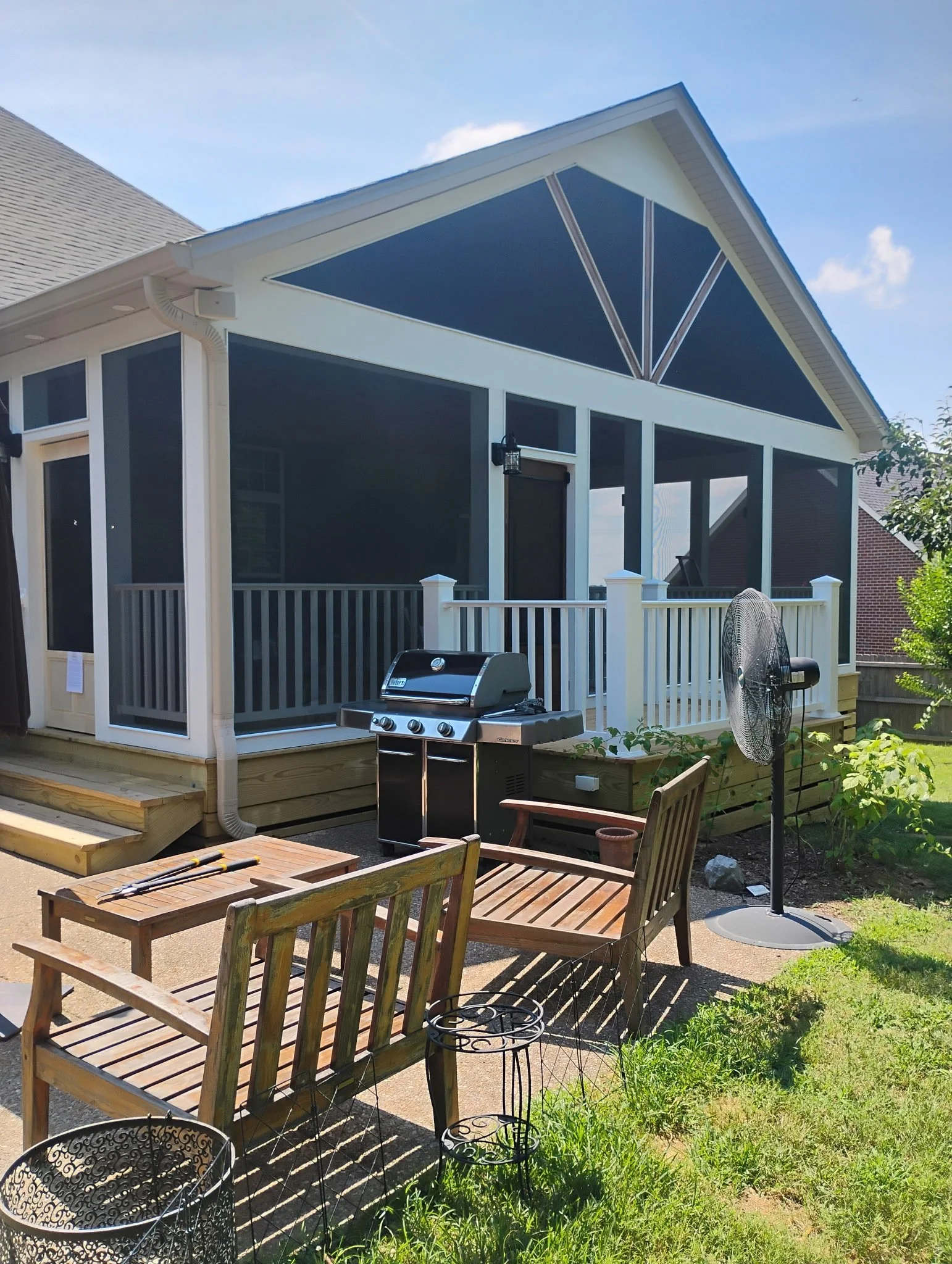 A backyard patio with wooden chairs, a grill, a standing fan, and a screened porch attached to a house with a blue sky above.