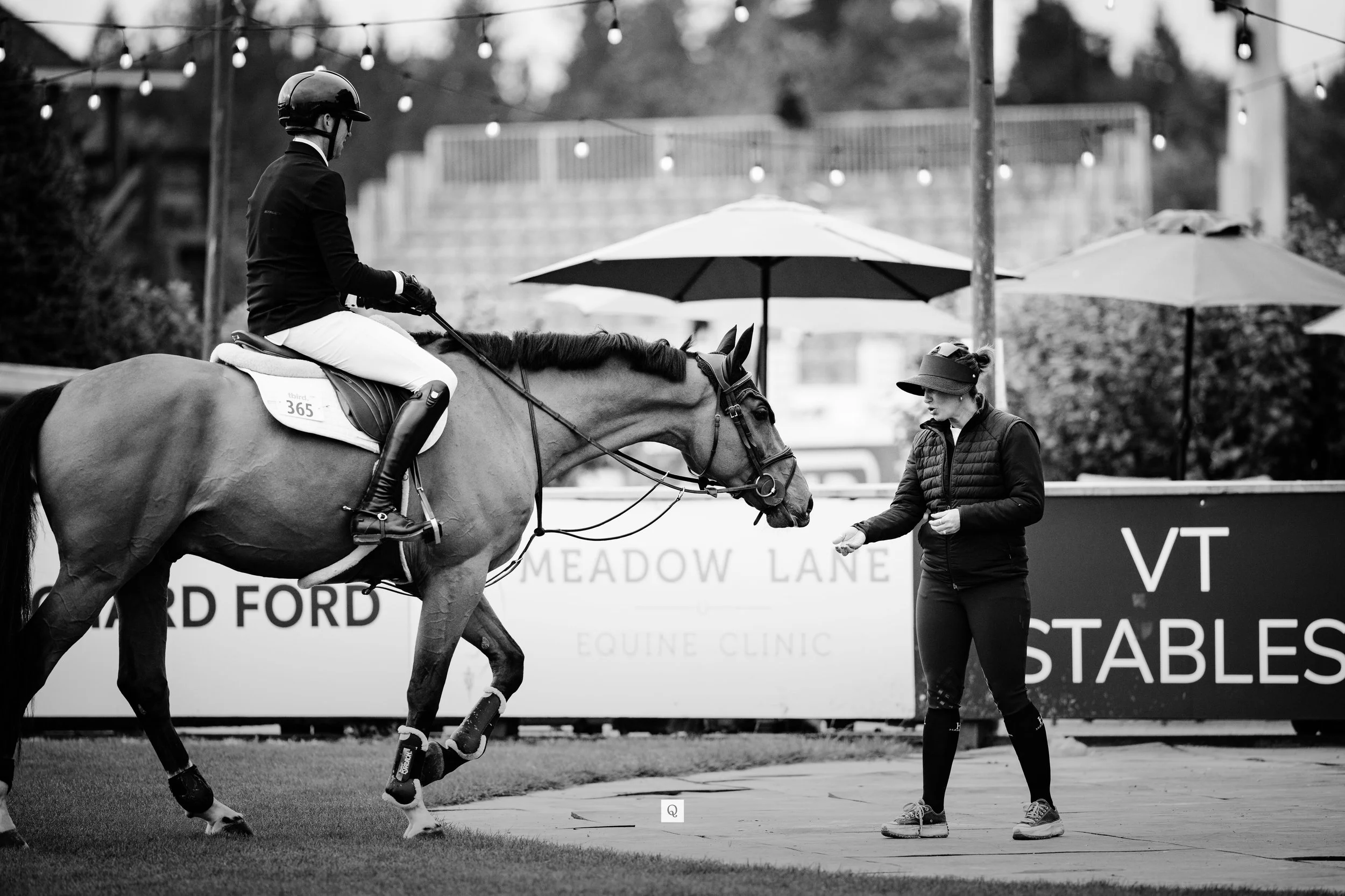 A person on horseback receiving instructions from a woman at an equestrian event, with signs and umbrellas in the background.