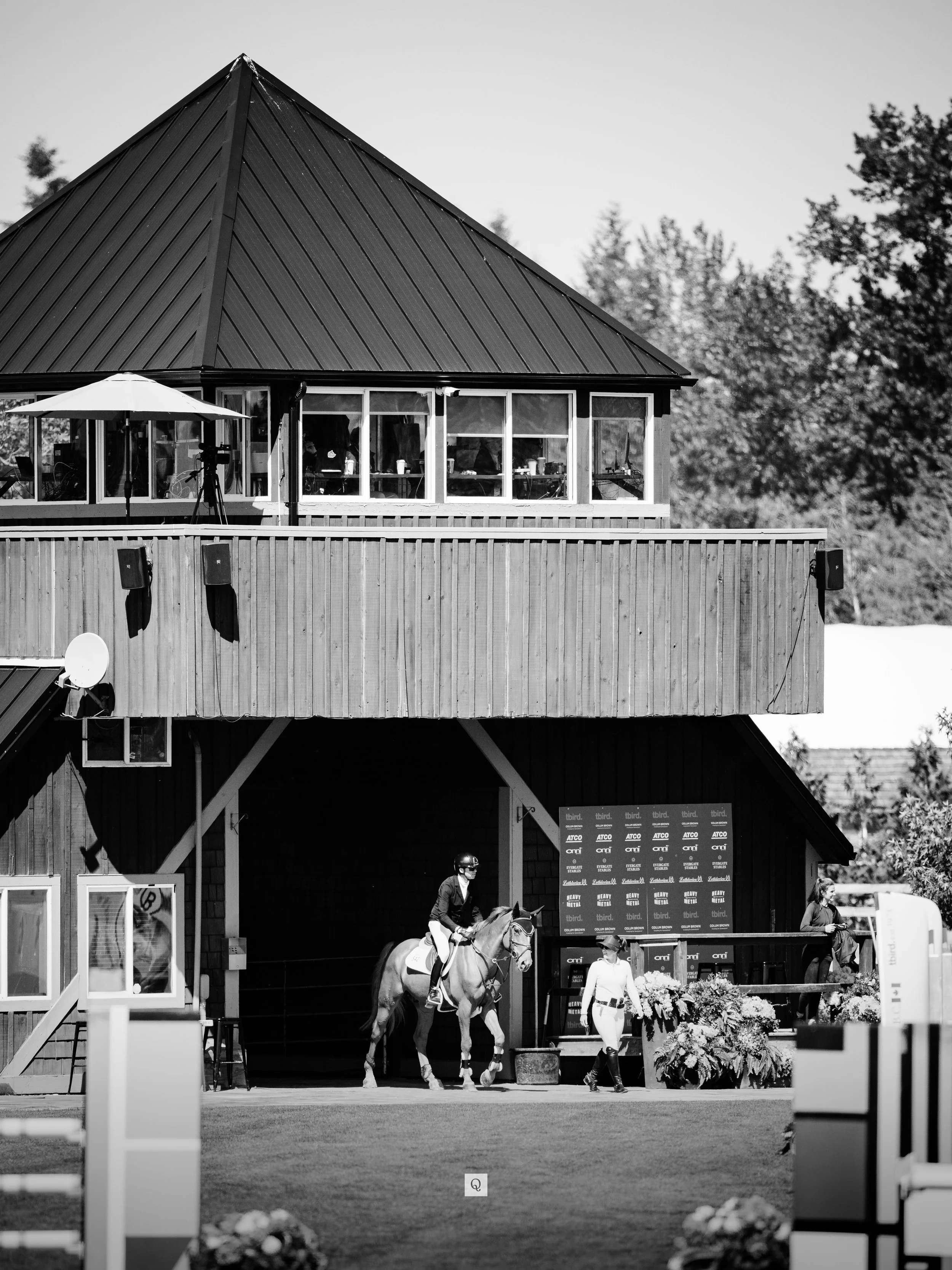 Black and white photo of an equestrian event with a rider on a horse in the foreground and a building with windows and a roof in the background. People are visible around the horse and on the balcony.