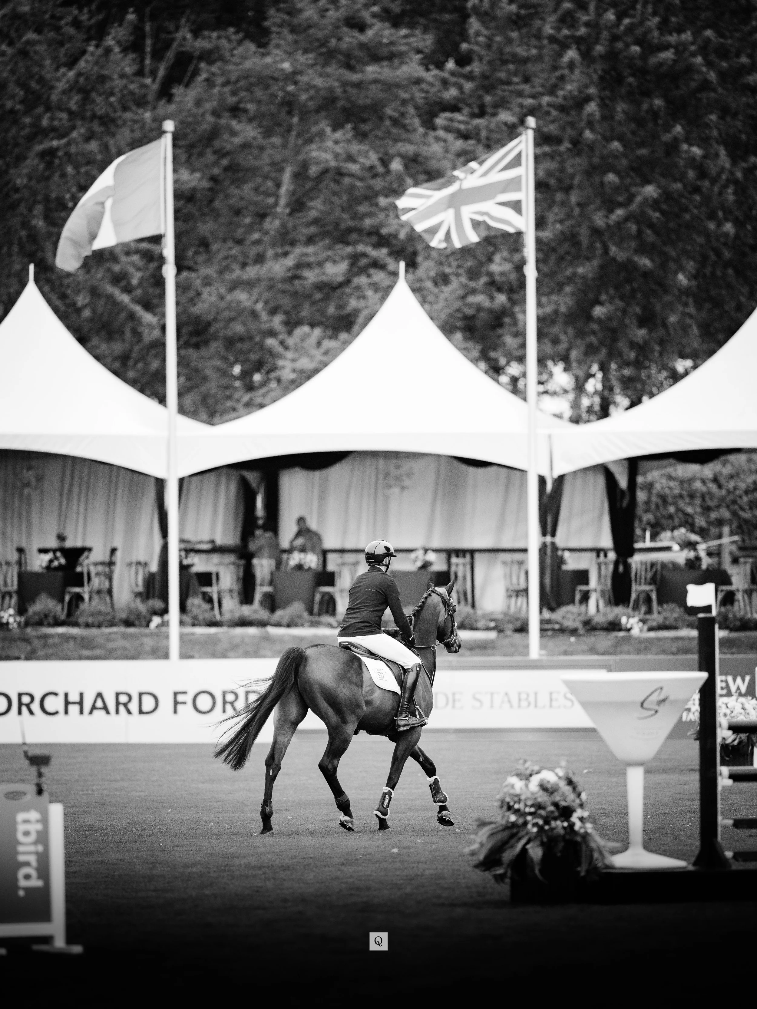 A person riding a horse in a competition, with antennas and flags on poles, including a Union Jack flag, in the background at an outdoor venue.