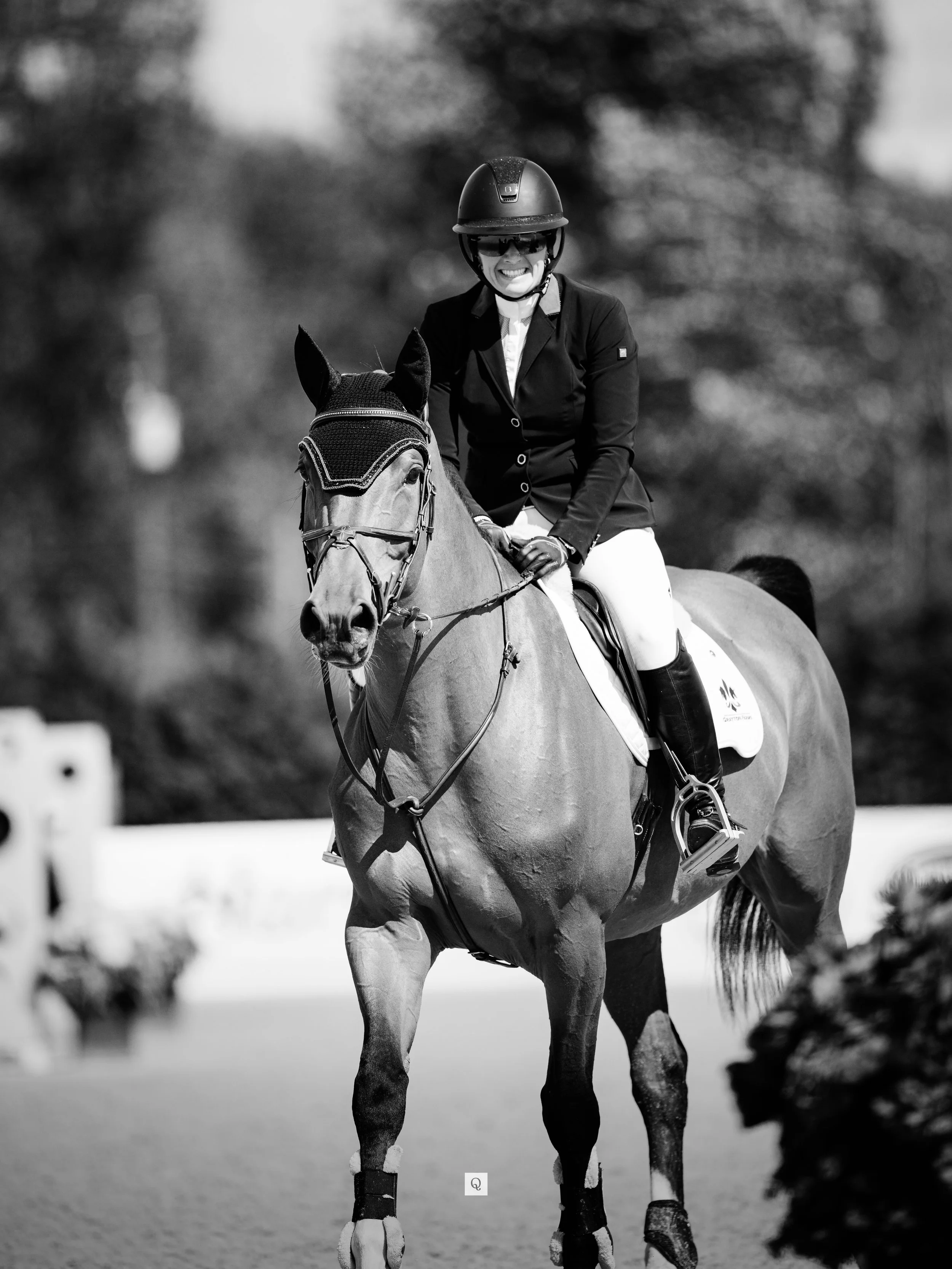 A woman riding a horse during a showjumping competition, smiling, wearing a helmet and riding attire, in an outdoor setting.
