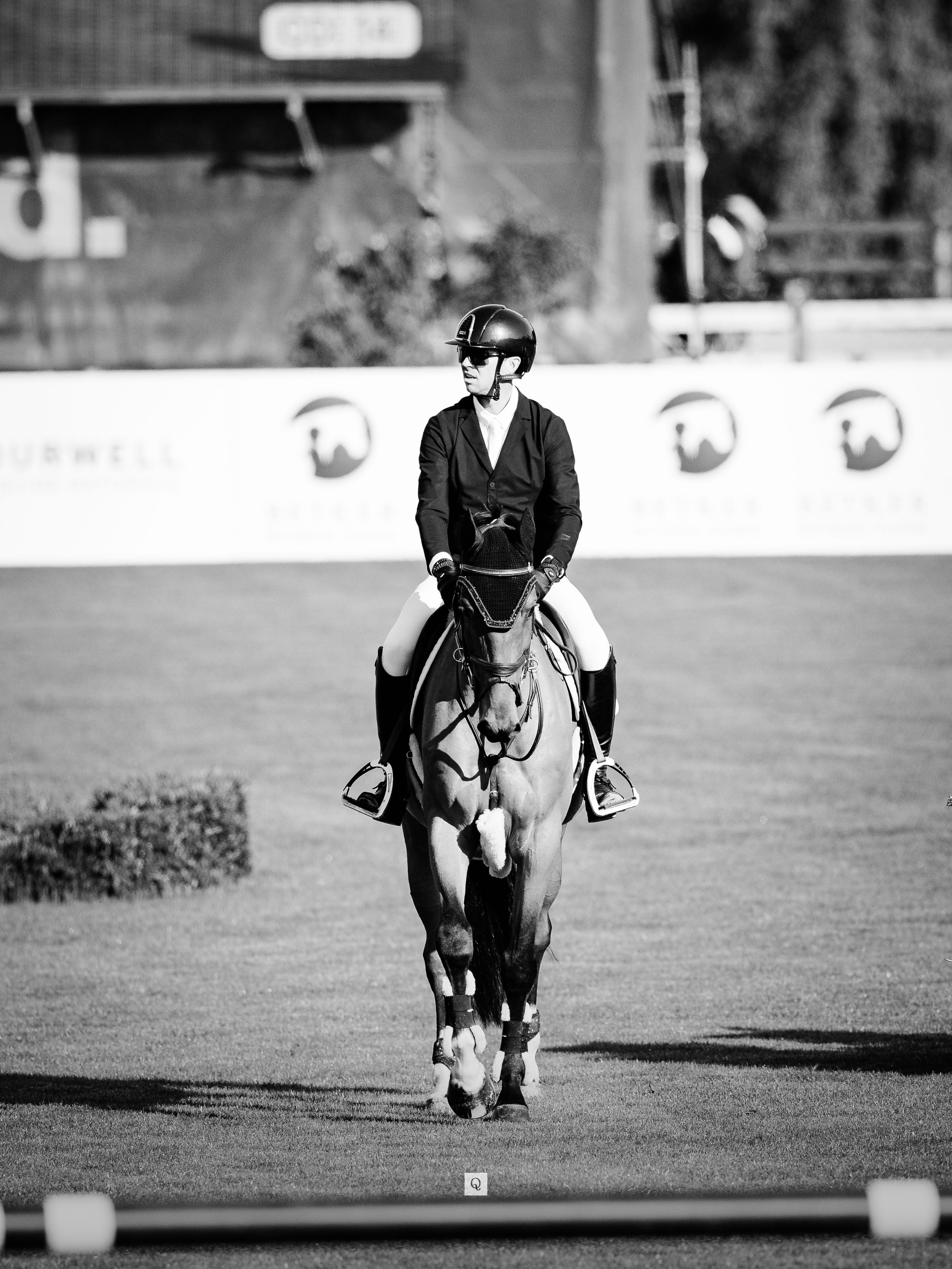 A person in equestrian riding attire, including a helmet, sunglasses, jacket, and riding boots, is riding a horse on a field.