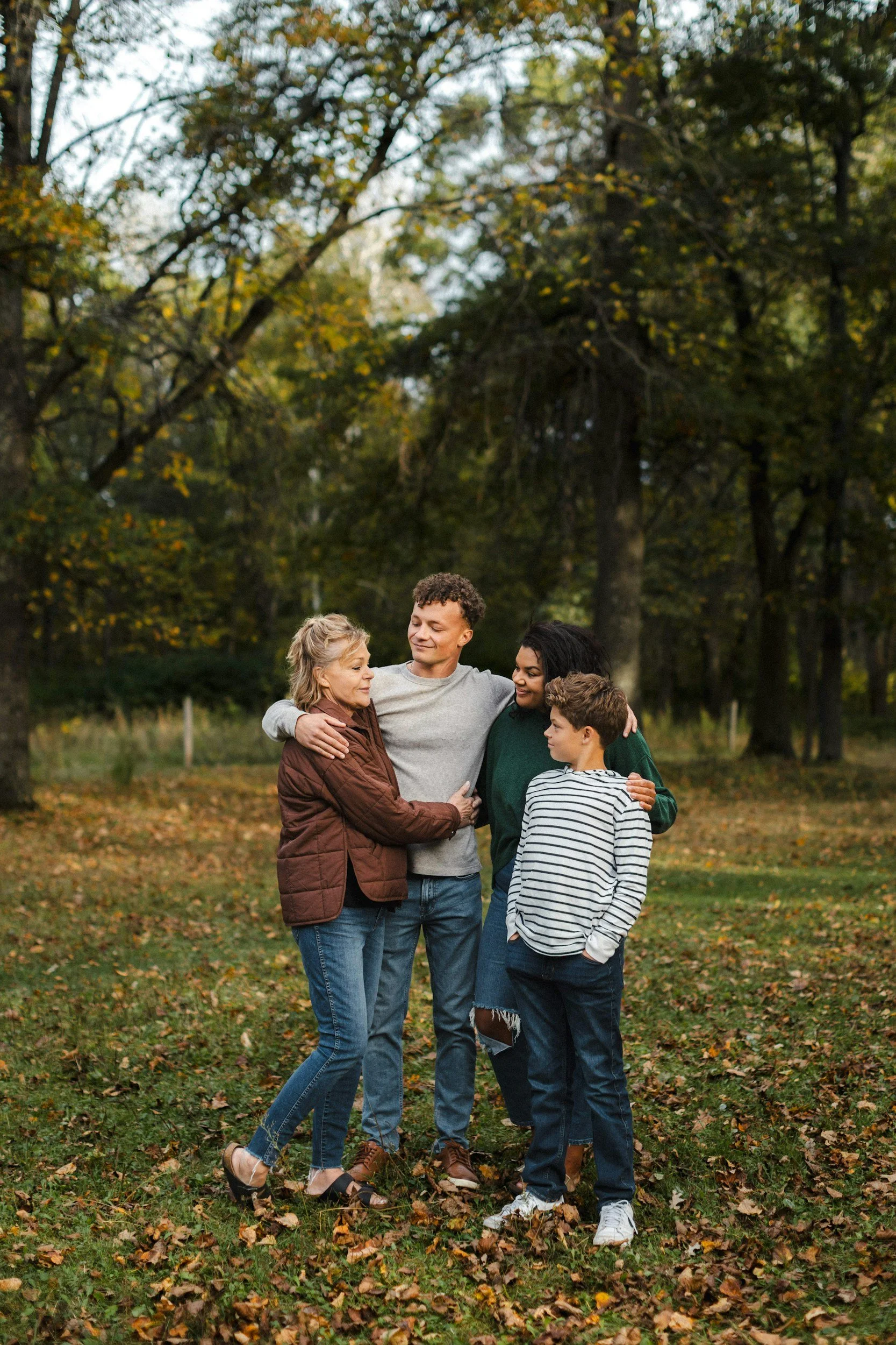 A multigenerational family standing outdoors in a park during fall, embracing and smiling at each other.