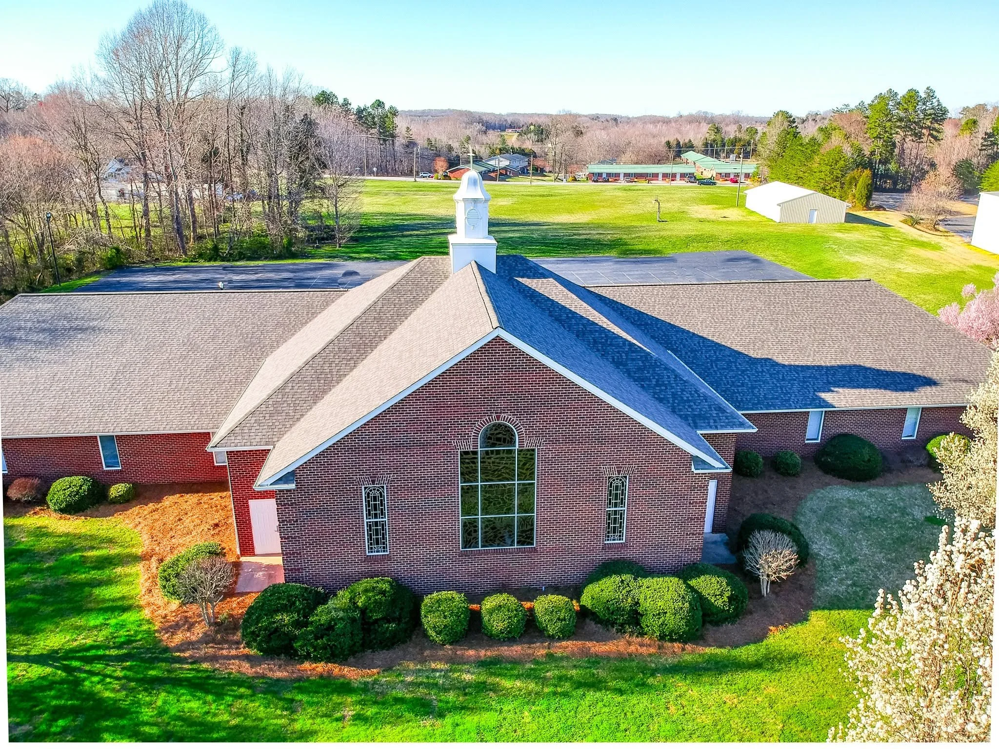 Aerial view of a red brick church with a gray shingled roof, white bell tower, large stained glass window, surrounded by neatly trimmed bushes and grass, with a parking lot and trees in the background.
