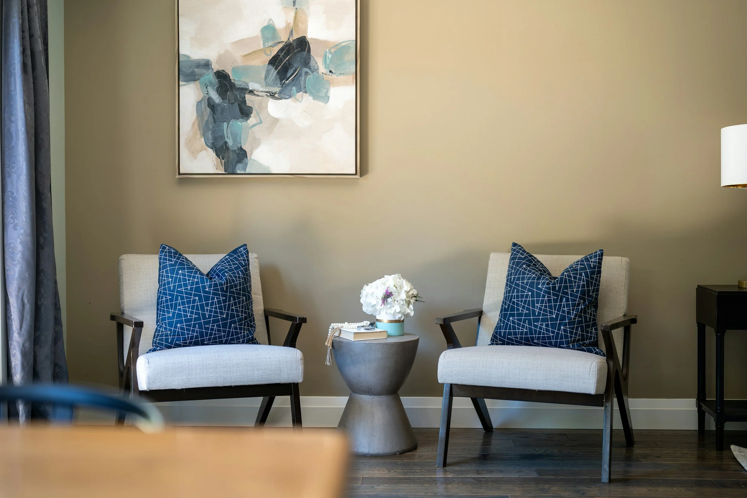 Living room with two white armchairs with blue geometric pillows, round gray side table with white flowers, abstract artwork on beige wall, cream curtains on left, white floor lamp on right.