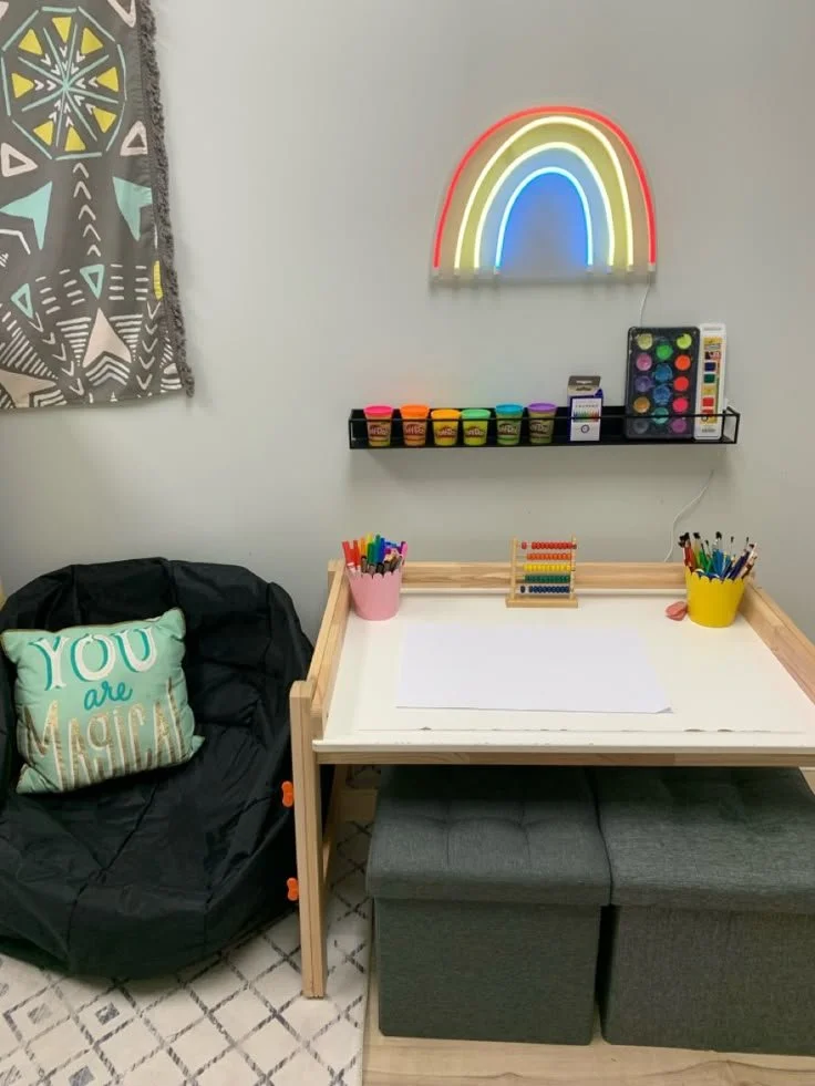 A cozy room corner with a black bean bag chair featuring a mint green and gold pillow that says "You are Magic." Next to it is a small table with art supplies, including markers, paint, and an abacus. On the wall is a colorful neon rainbow sign and a shelf with paint pots and a watercolor set. There is also a patterned fabric wall hanging and a small cushioned bench under the table.