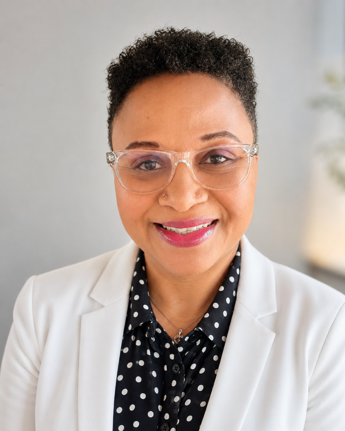 Portrait of a woman wearing clear glasses, a white blazer, and a black polka dot blouse, smiling indoors with a light background.