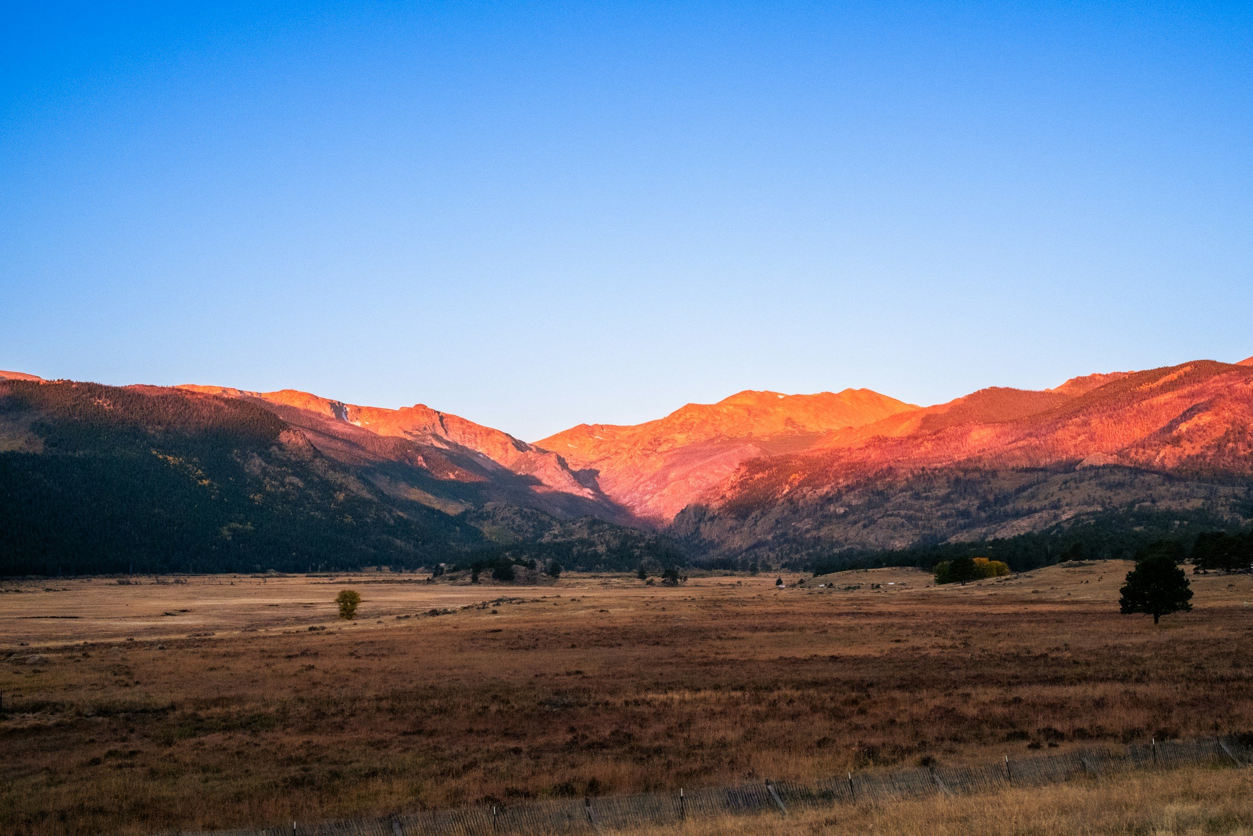 Scenic landscape of distant mountains illuminated with warm sunlight, with a flat plains foreground and a few sparse trees.