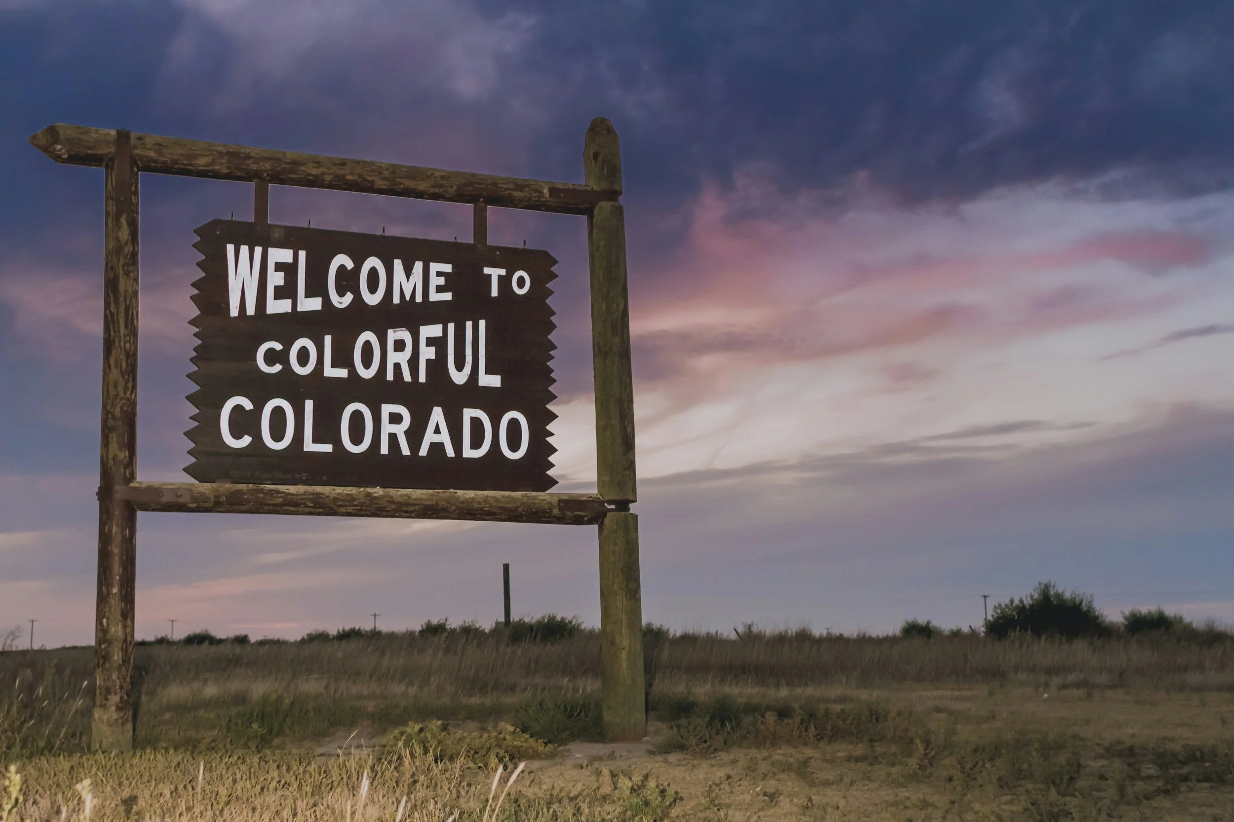 A weathered wooden sign reading 'Welcome to Colorful Colorado' against a cloudy sky at dusk.
