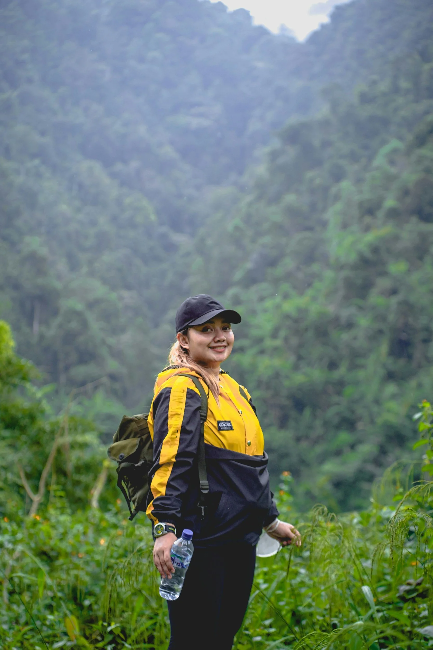 A woman in outdoor gear holding a water bottle and a hat, smiling in a lush green forest.