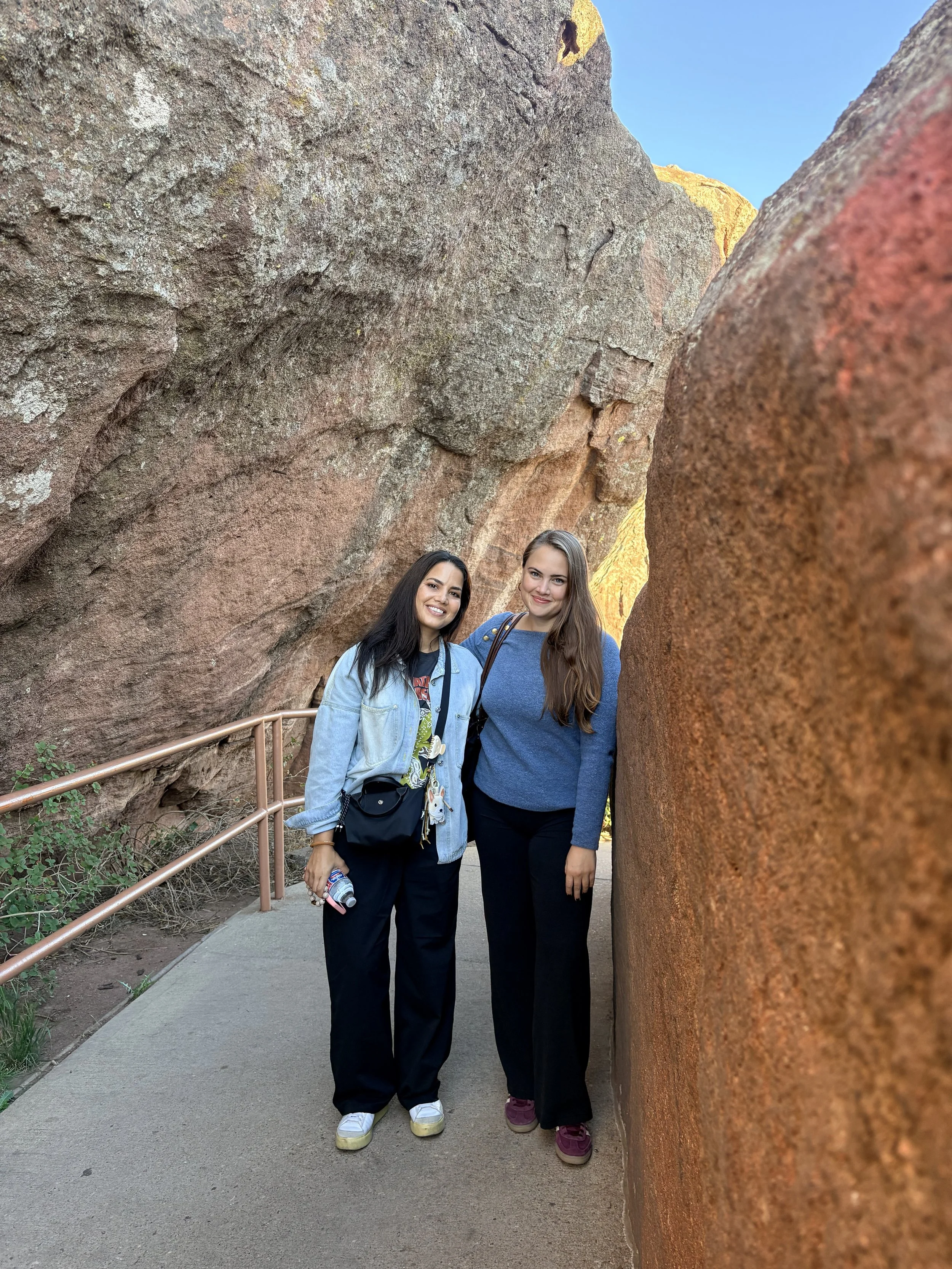 Two women standing close together outdoors between large rock formations, smiling at the camera. One woman has long dark hair and is wearing a light denim jacket, black pants, and white sneakers. The other woman has long brown hair and is dressed in 