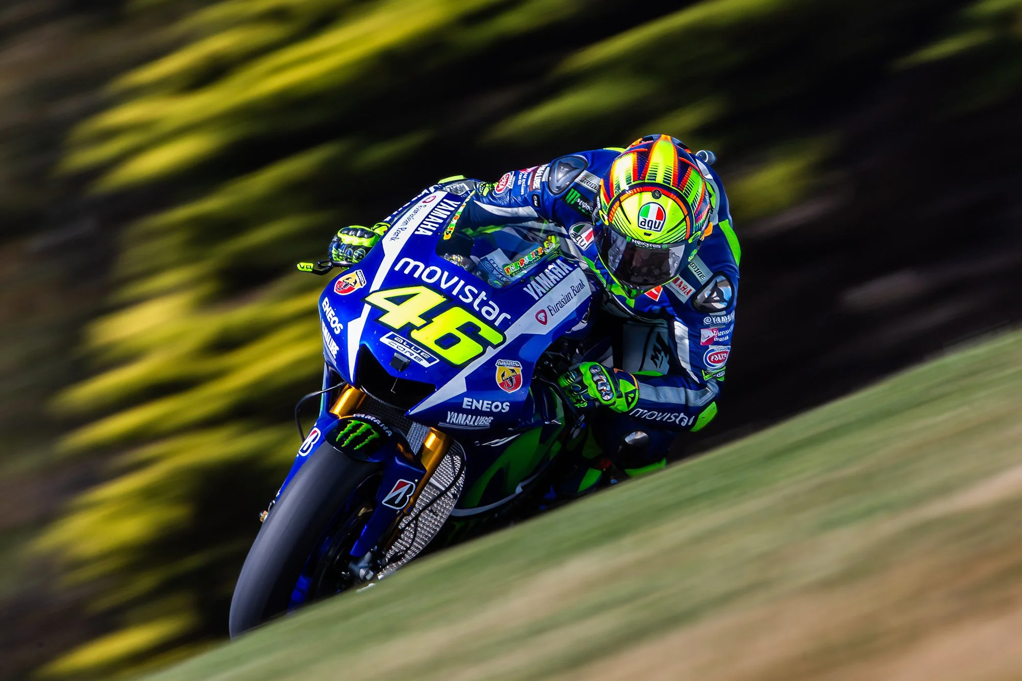 A motorcycle racer in a blue and green racing suit and helmet riding a blue Yamaha motorcycle on a race track with a blurred background.
