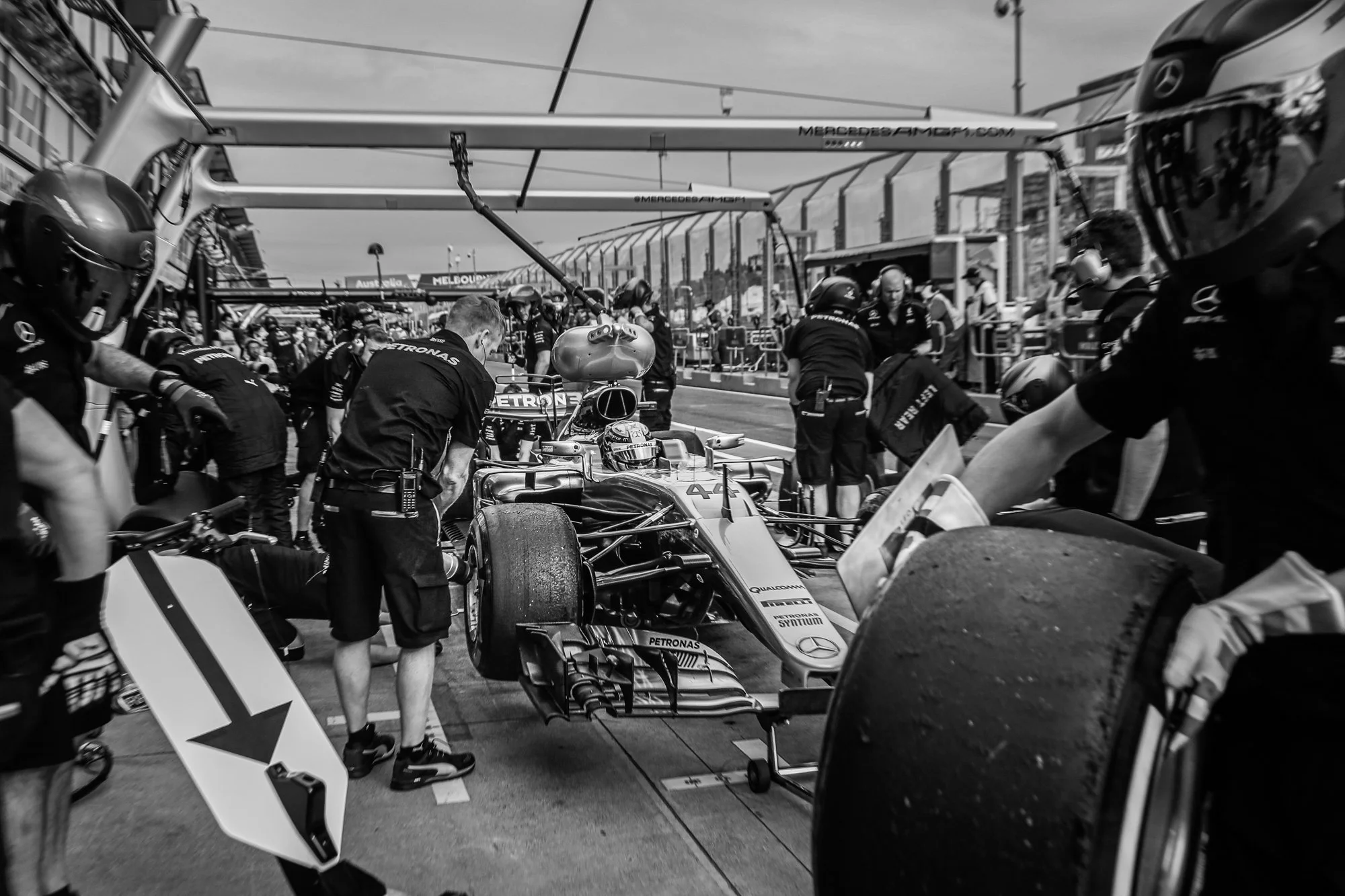 Race car team members working on a Formula 1 car in the pit lane, with team helmets and tools visible, Mercedes branding on the car.