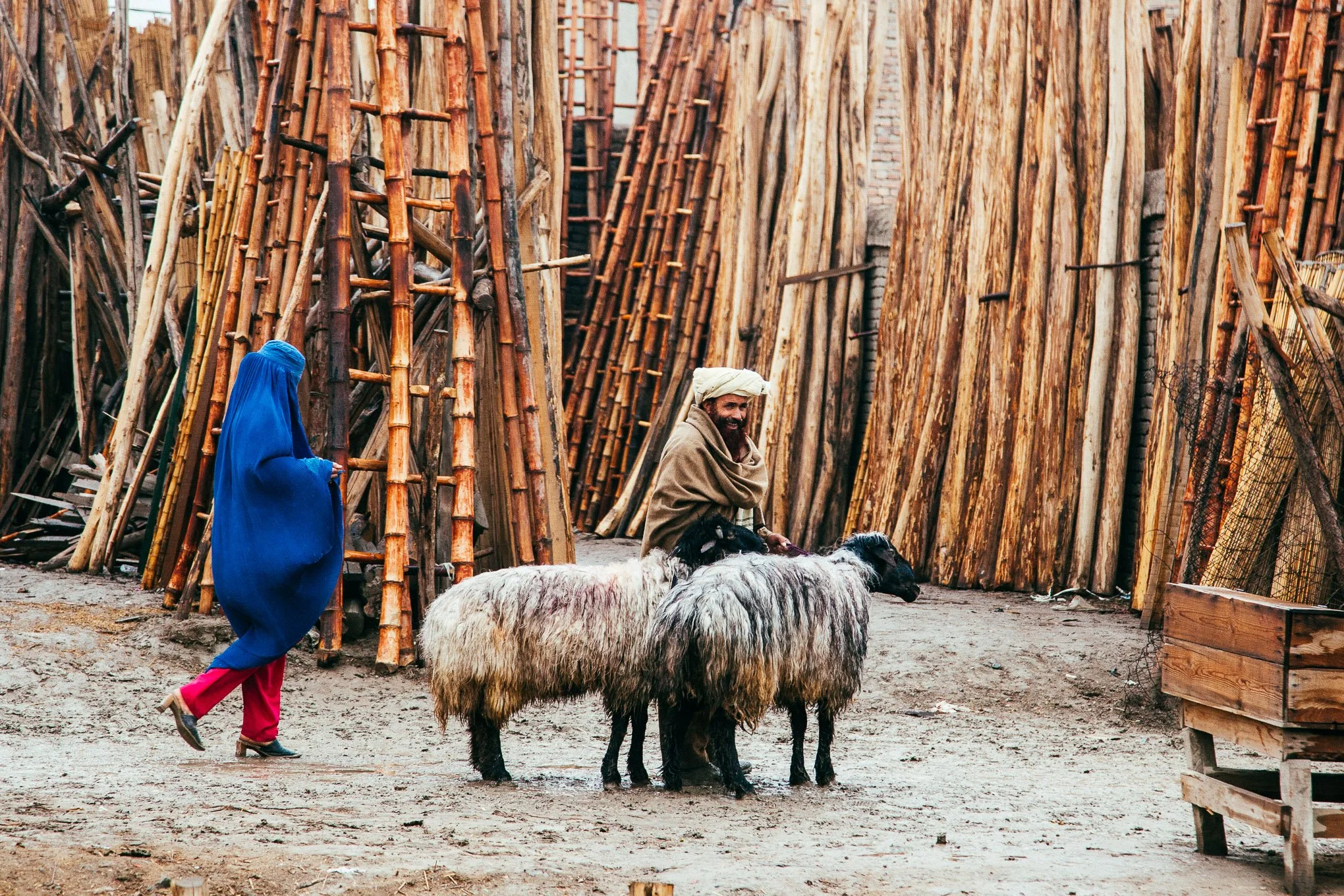 An Afghan man with two sheep smiles as a woman in a blue burqa walks past. 