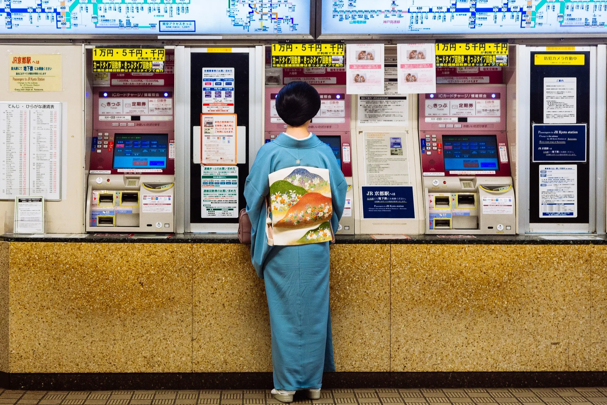A woman wearing a traditional blue kimono with floral patterns, standing at a ticket vending machine in a train station in Japan.