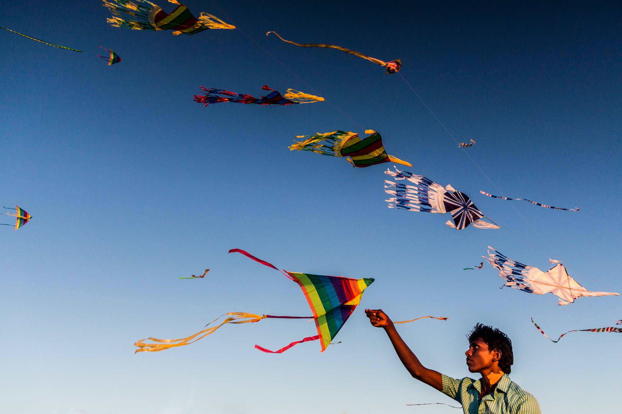 A young man pulls a kite string as he flies his kite as he is surrounded by other flying kites as the sun sets on Galle Face