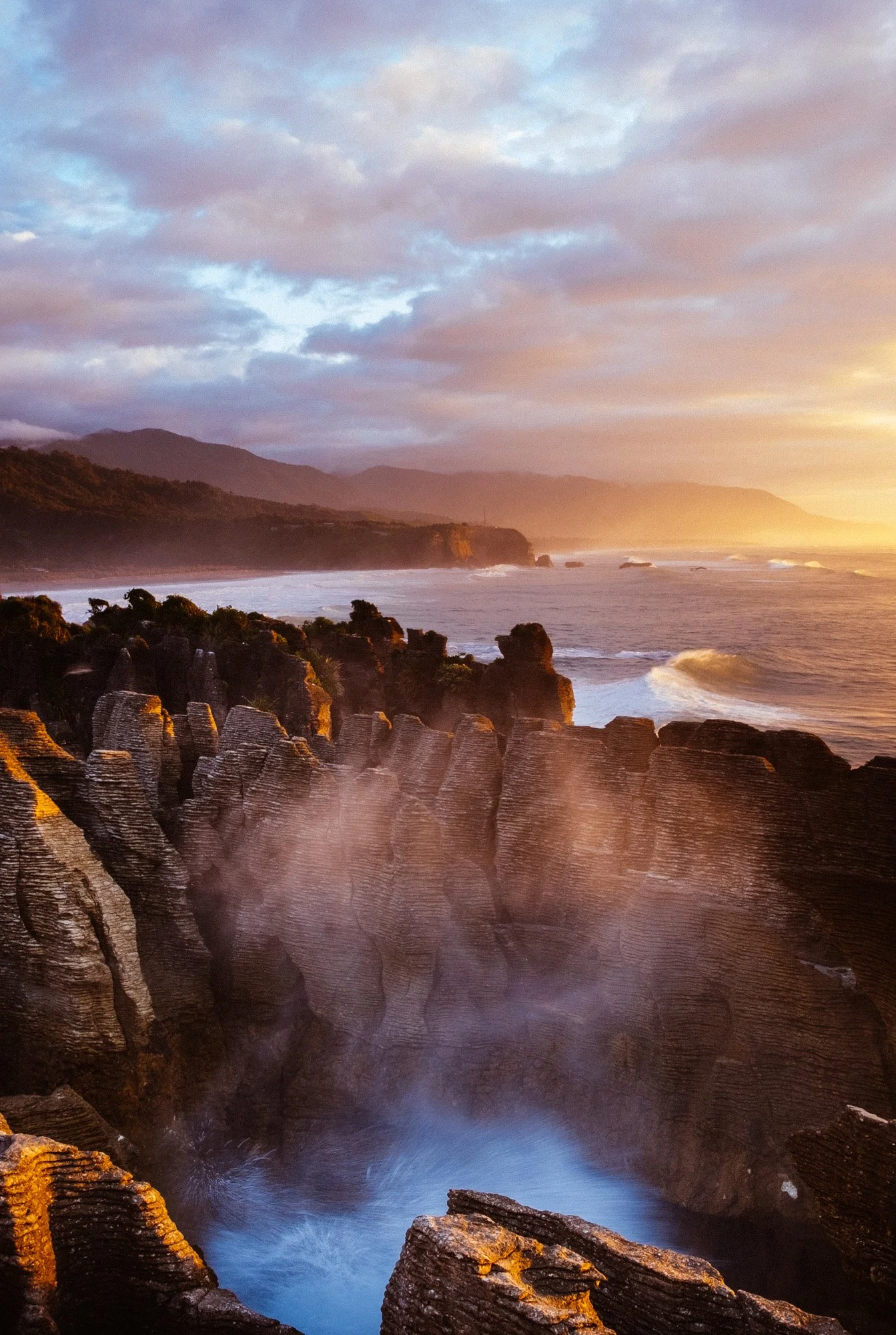 Sunset over a rocky coastal landscape with mountains in the background and ocean waves crashing against the rocks.