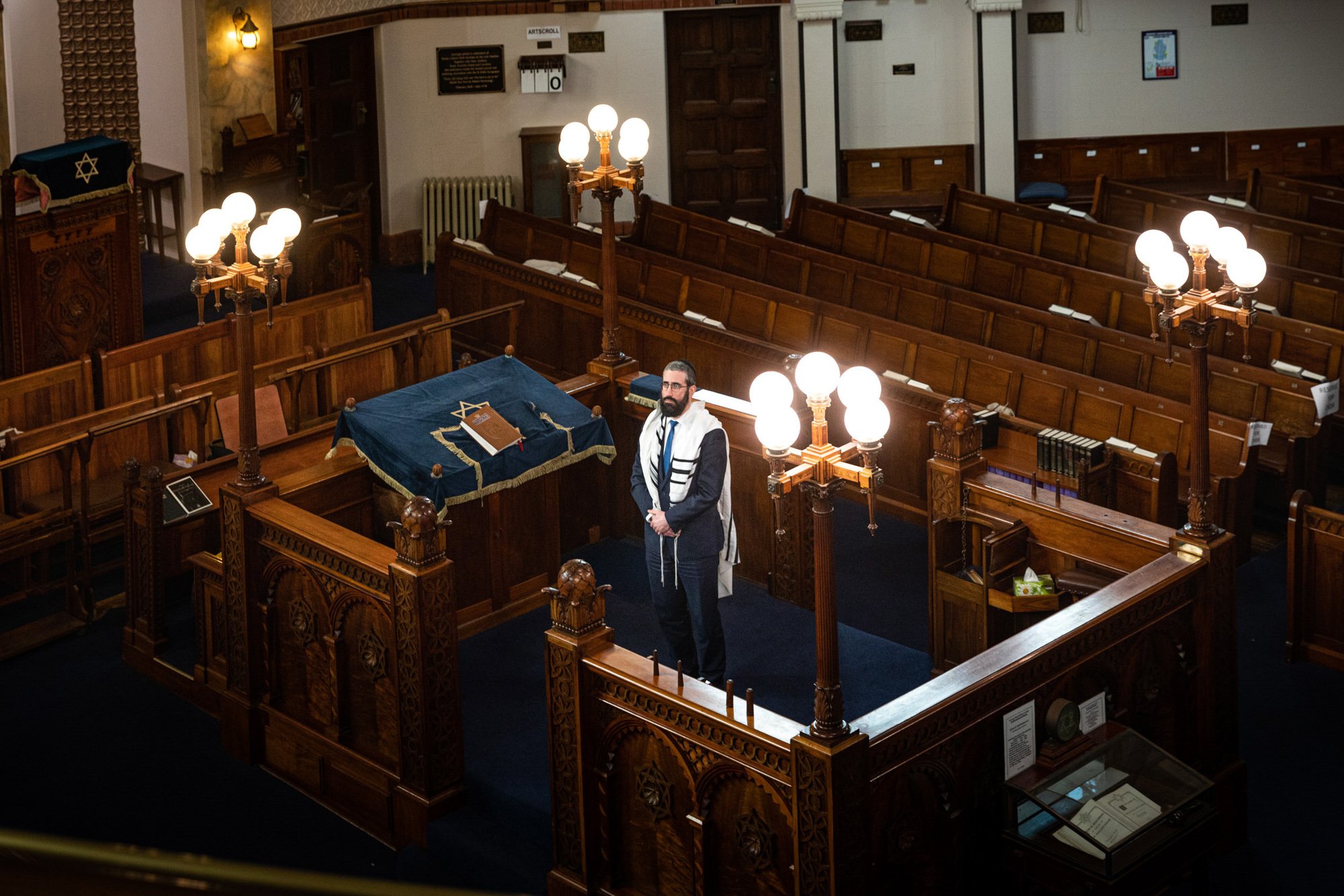 Interior of a synagogue with wooden pews, ornate traditional woodwork, and lit brass chandeliers. A person in formal attire and tefillin stands near the front, with a Torah covered in blue velvet and gold embroidery on a table.