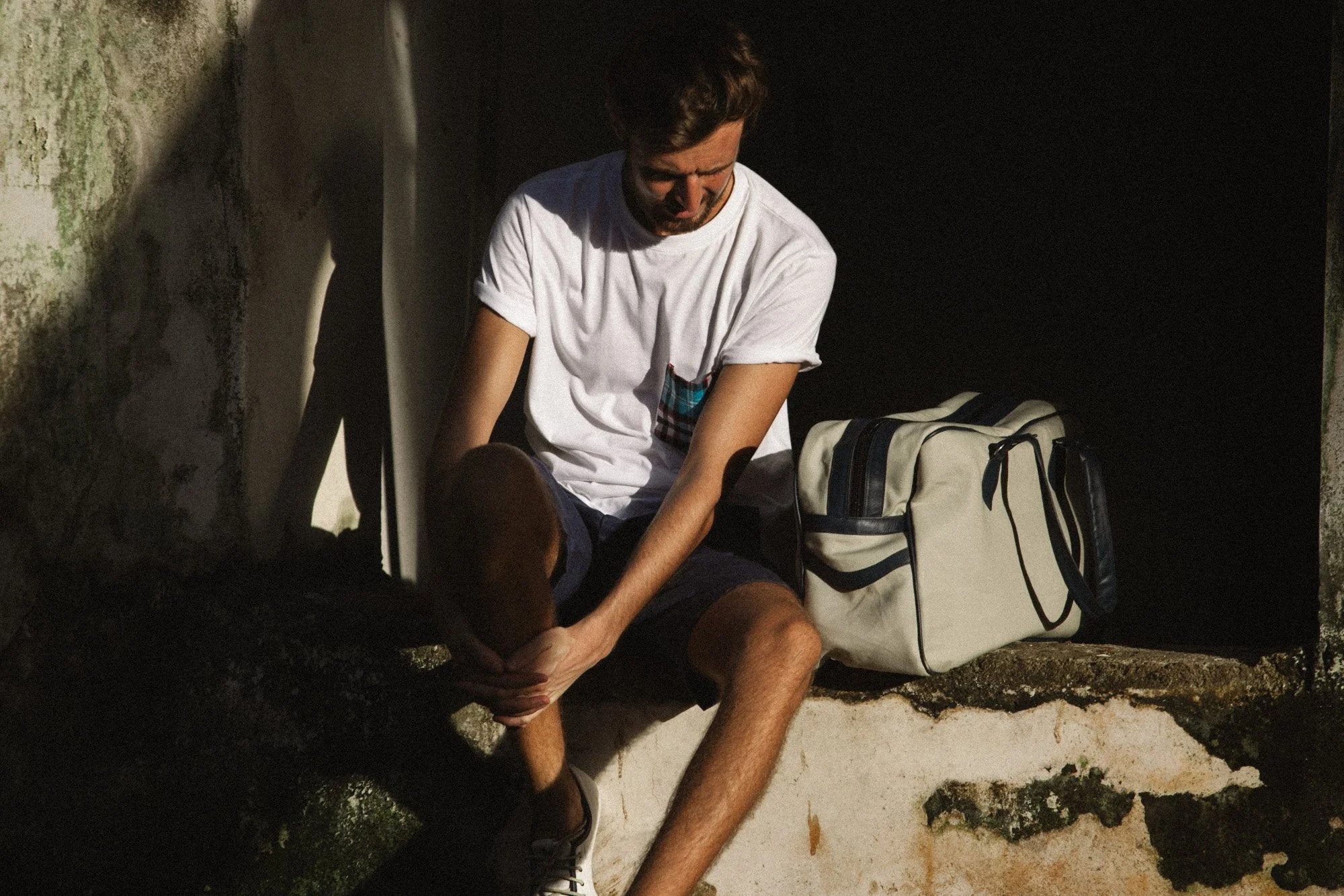 A man ties his shoe beside a travel bag in warm window light during a lifestyle fashion shoot.