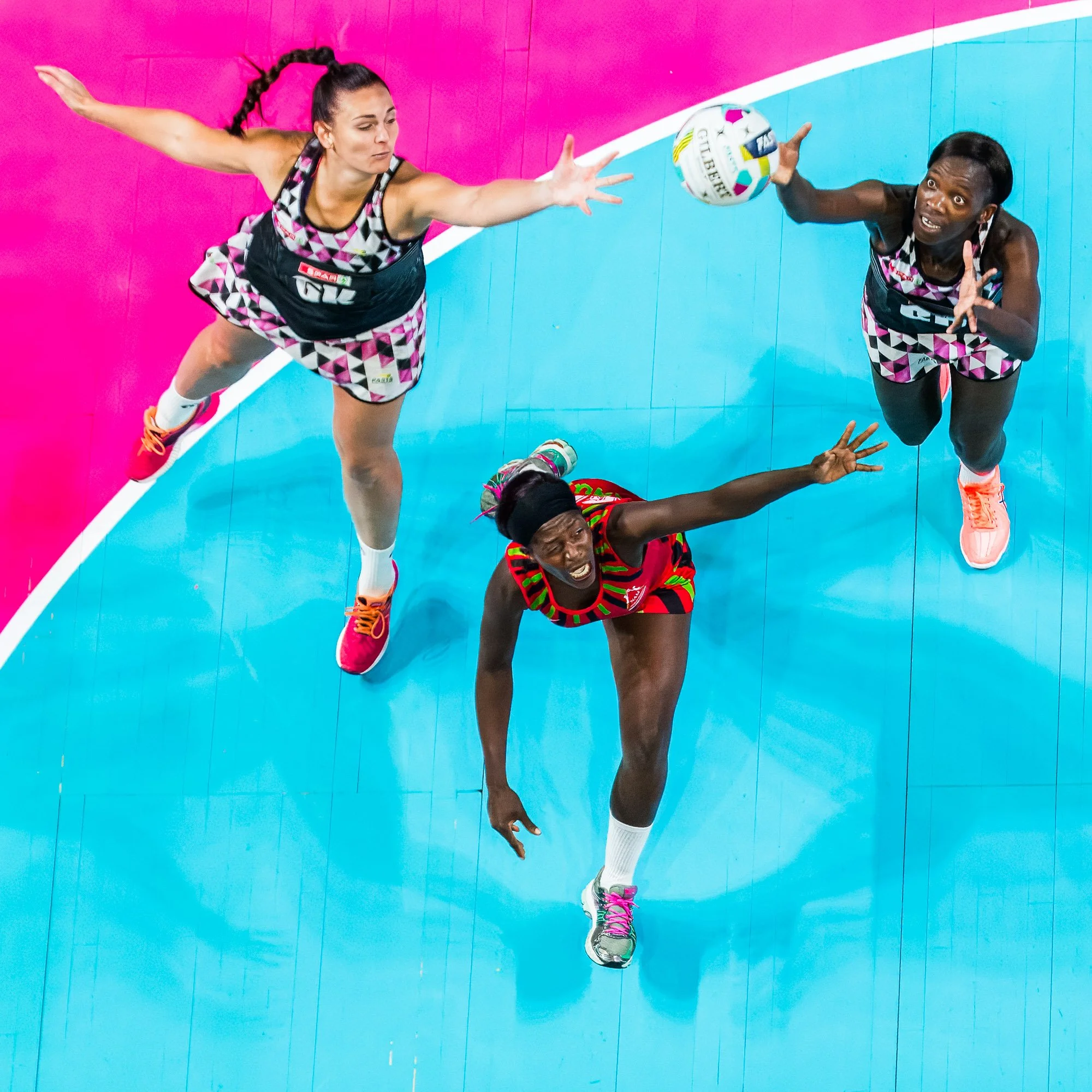 Two volleyball players in black and pink uniforms diving to save the ball during a match on a colorful court.