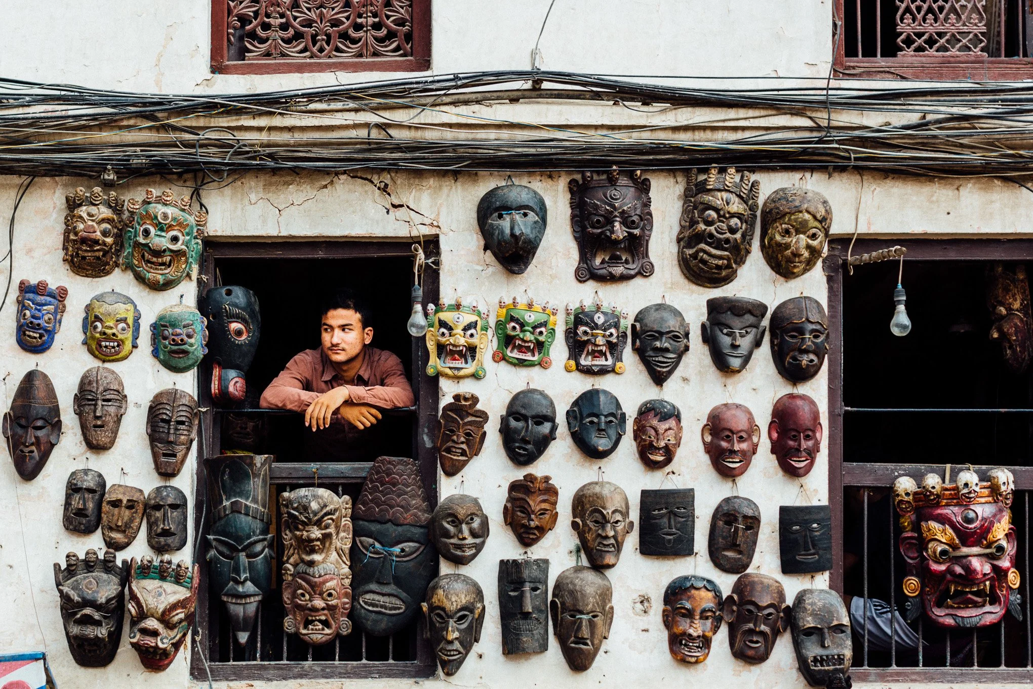A mask seller looks from a balcony above wooden carvings in Kathmandu Durbar Square.