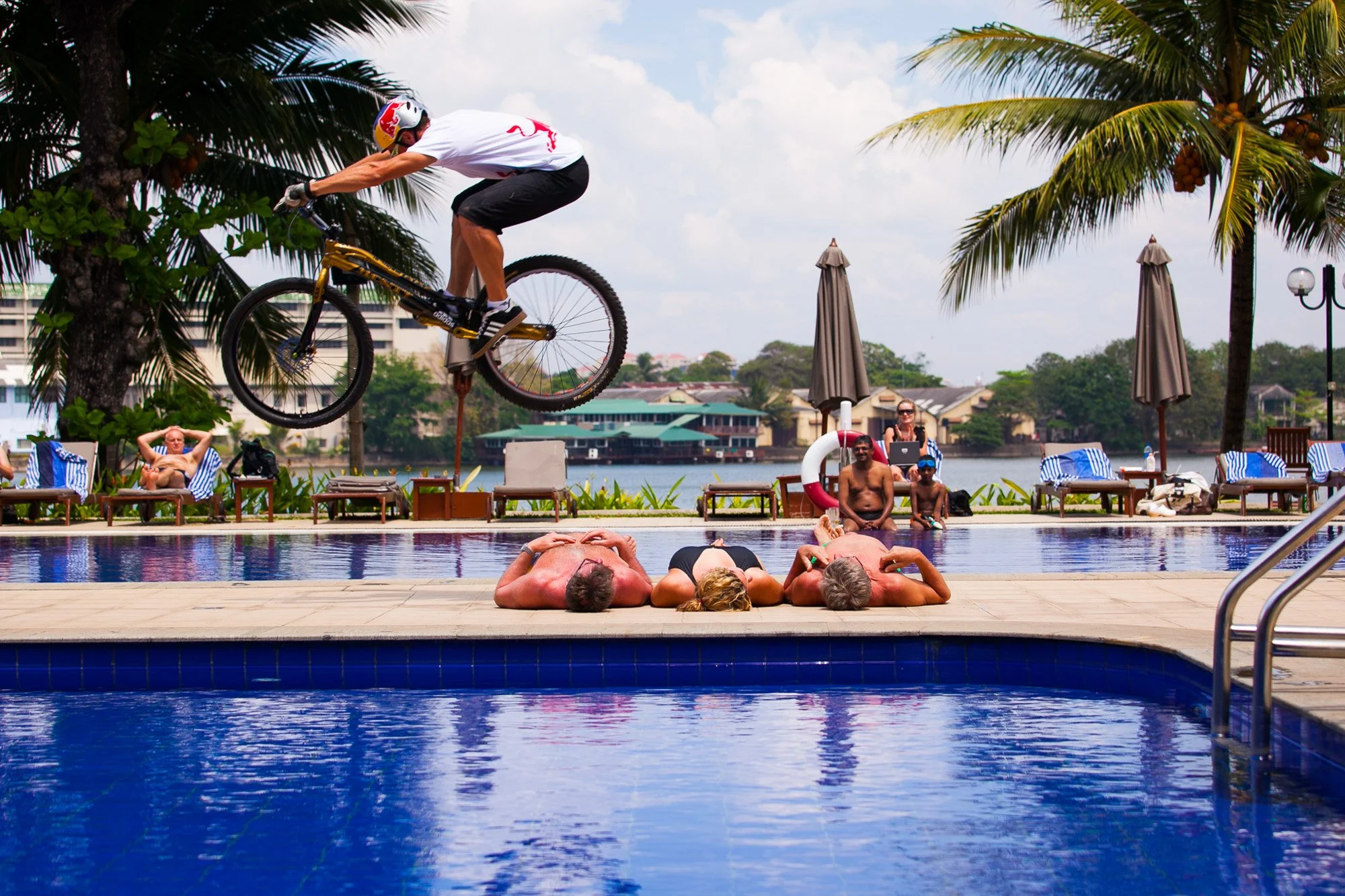 A person performs a bicycle stunt over a swimming pool, mid-air above reclining sunbathers, with palm trees and parasols in the background.