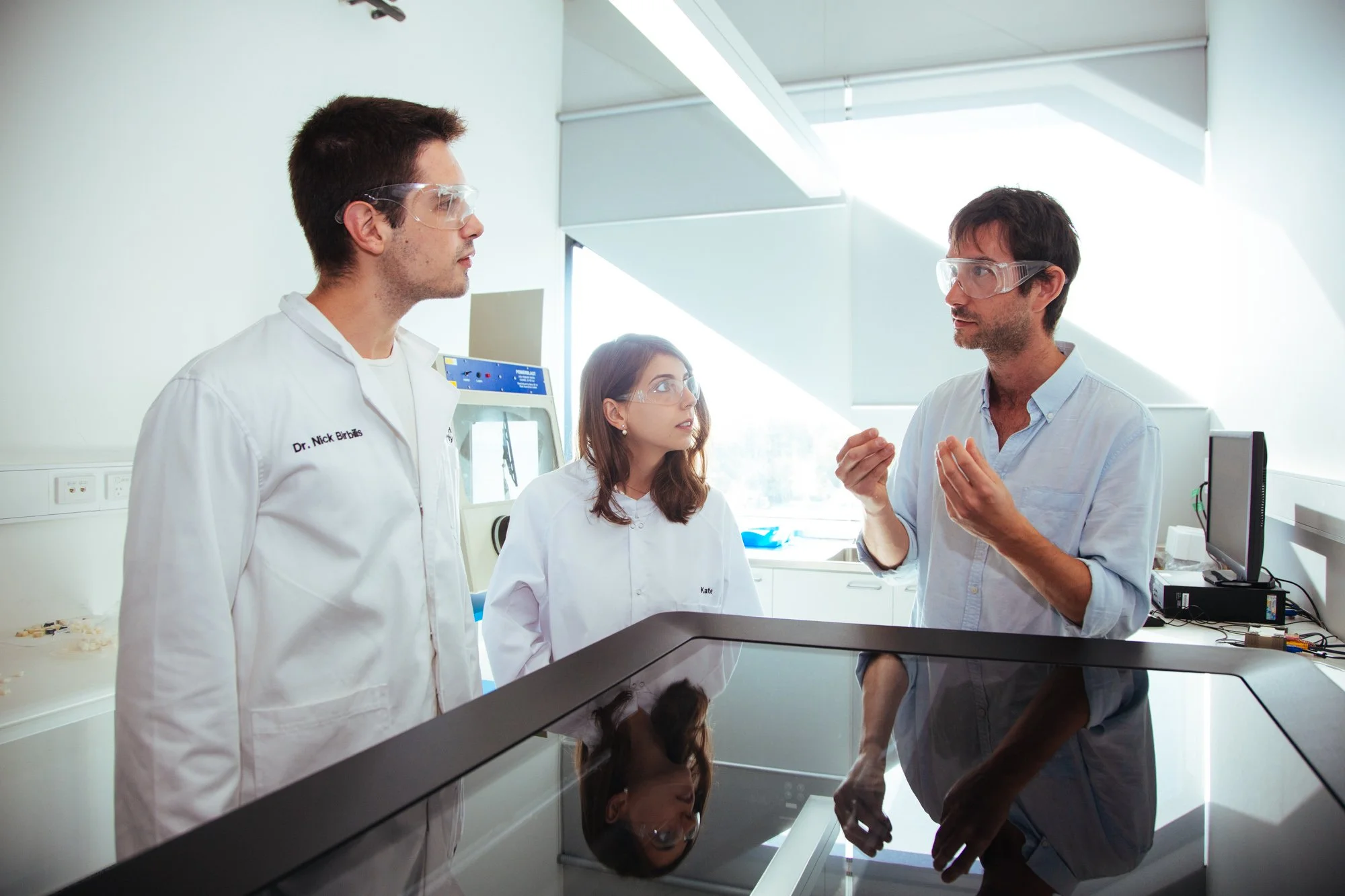Researchers in lab coats speak inside a university laboratory.
