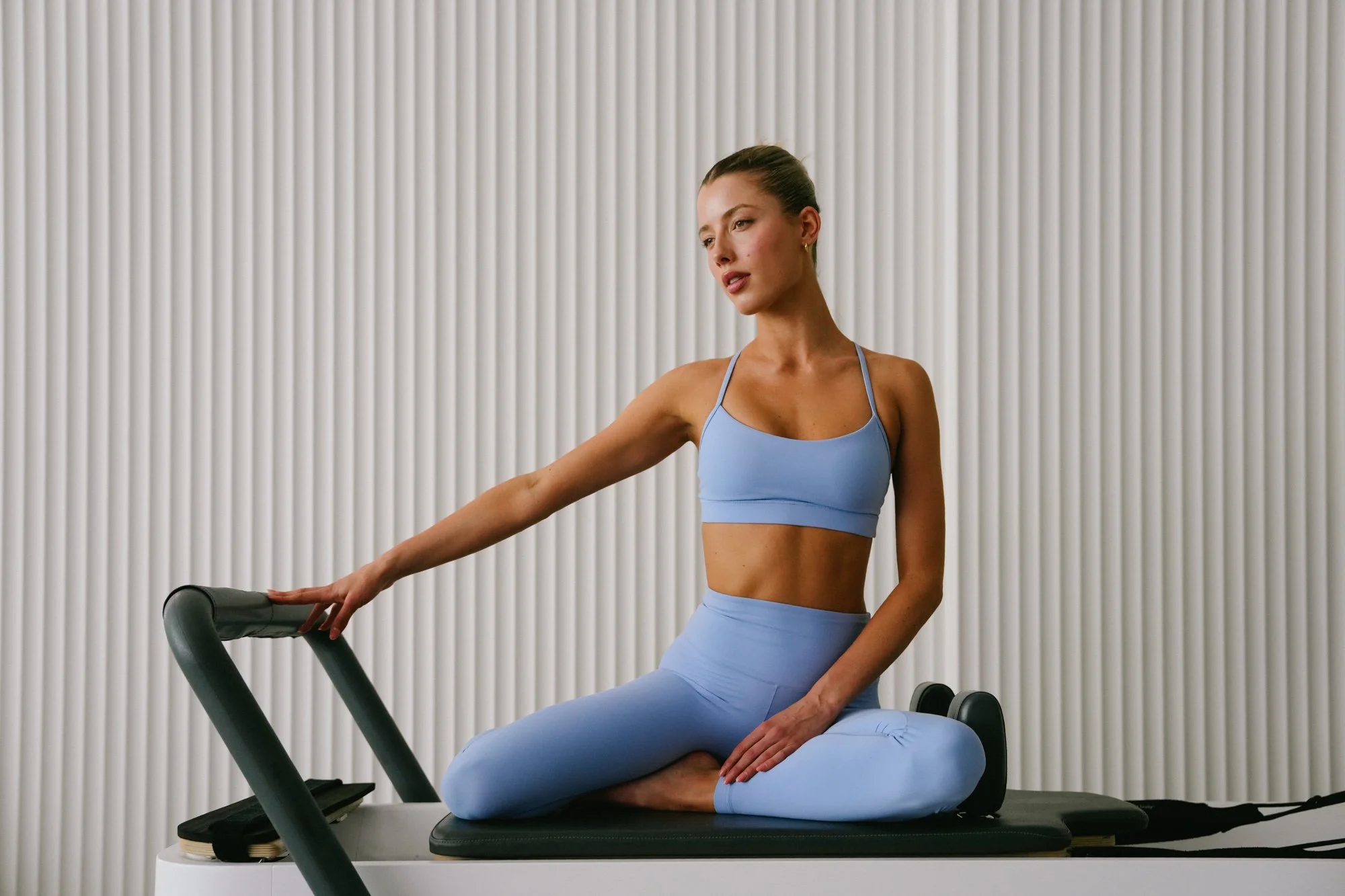 A woman poses on a pilates reformer while wearing blue activewear.