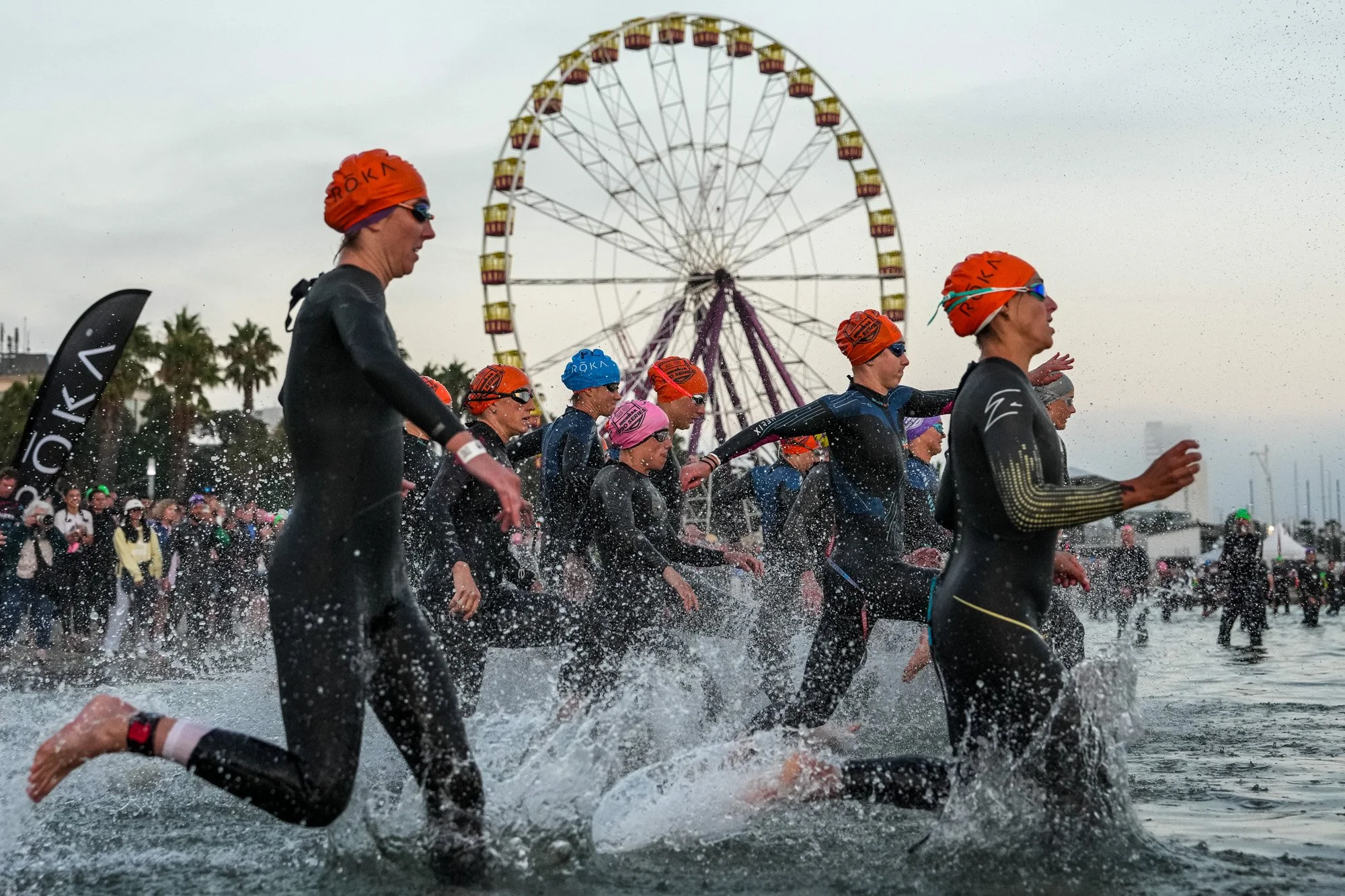A group of athletes, dressed in wetsuits and orange swim caps, running into the ocean during a triathlon event, with a Ferris wheel and spectators in the background.