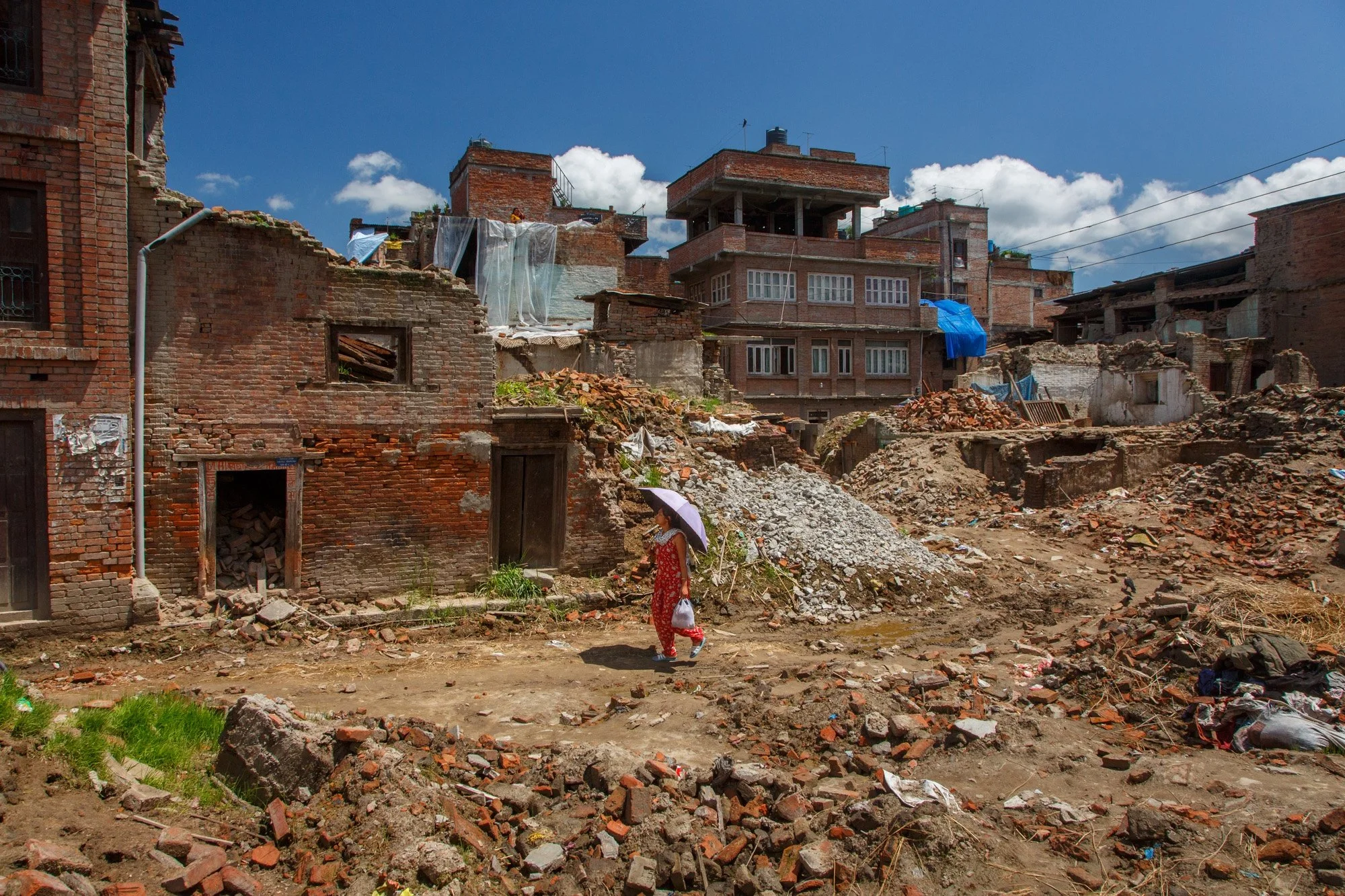A woman holding an umbrella walks through rubble and damaged buildings in Bhaktapur after the 2015 Nepal earthquake.
