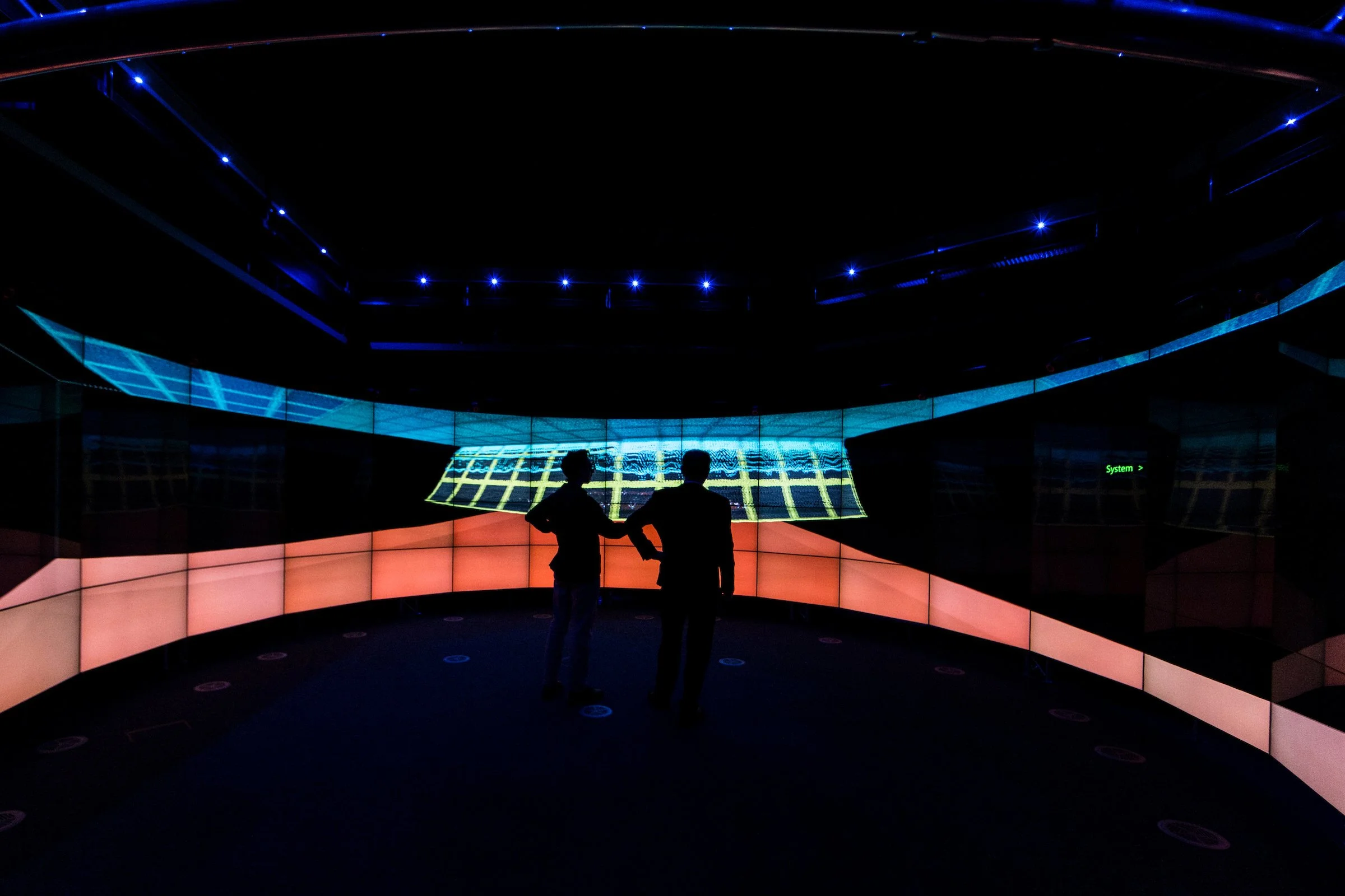 Two people stand inside a dark university visualisation room surrounded by illuminated screens.
