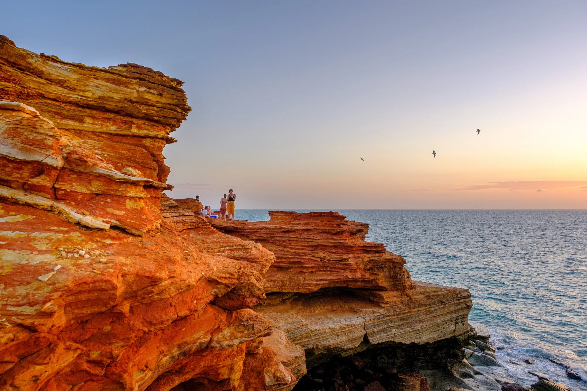 People sit along the red cliff rocks at Sunset looking out towards the Indian Ocean at Gantheaume Point in Broome
