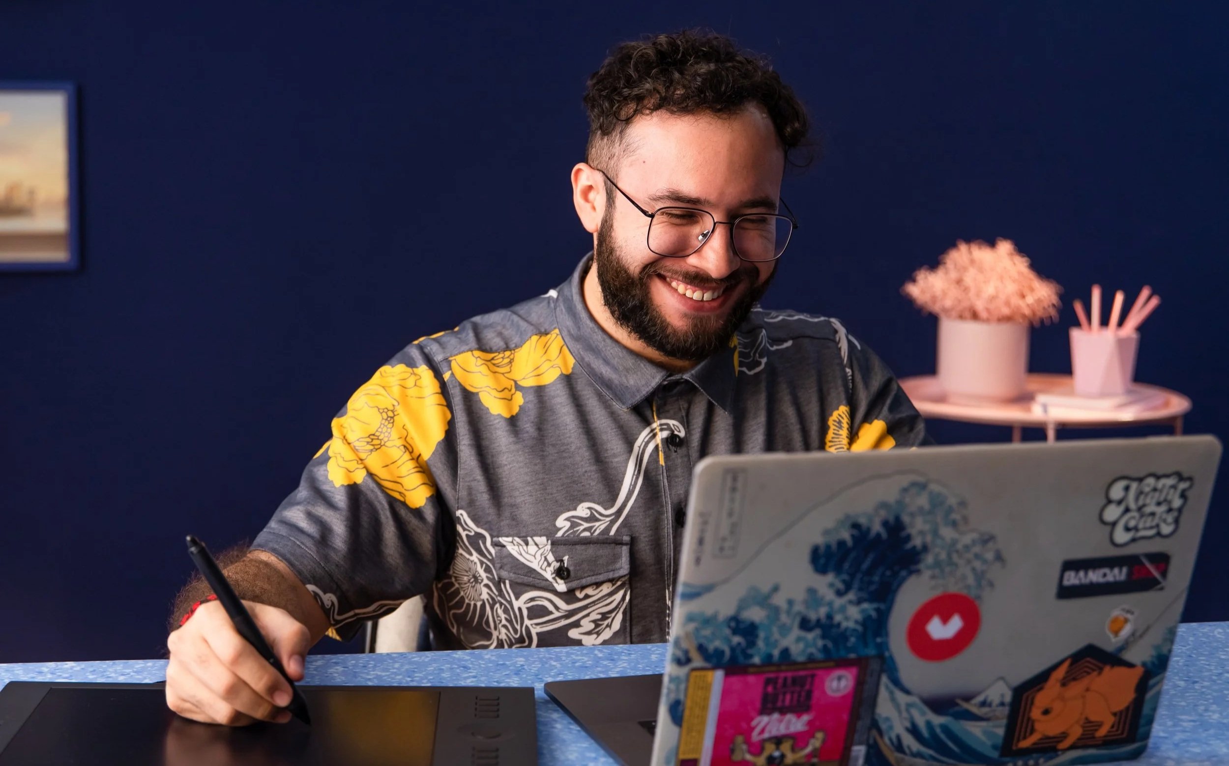 A young man with curly hair, glasses, and a beard, smiling while working on a laptop covered in stickers, sitting at a blue table against a dark blue wall, with pink flower decorations in the background.