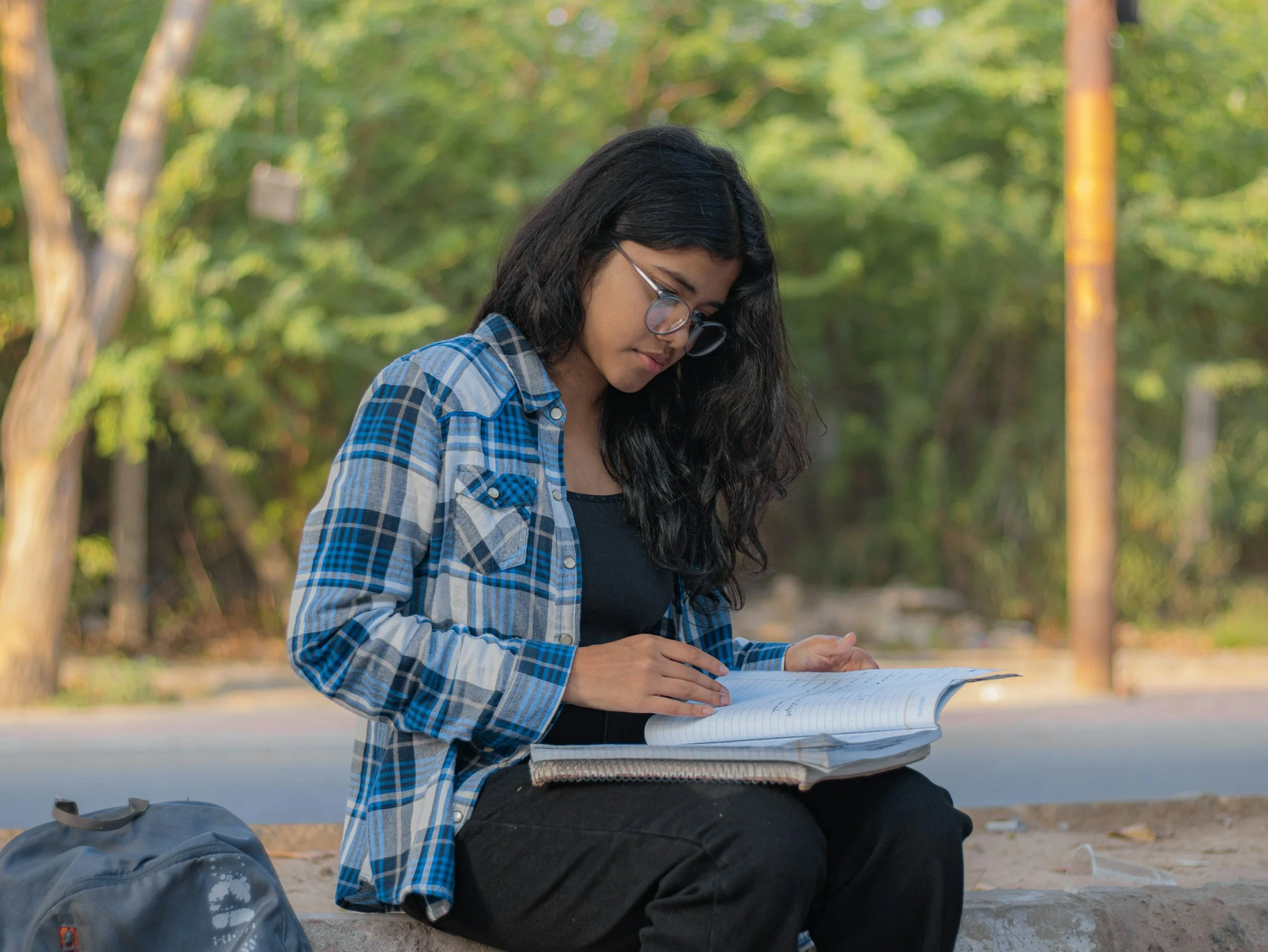 Young woman with long dark hair and glasses, wearing a blue plaid shirt, sitting outdoors at a park or forest, reading a book with trees in the background.