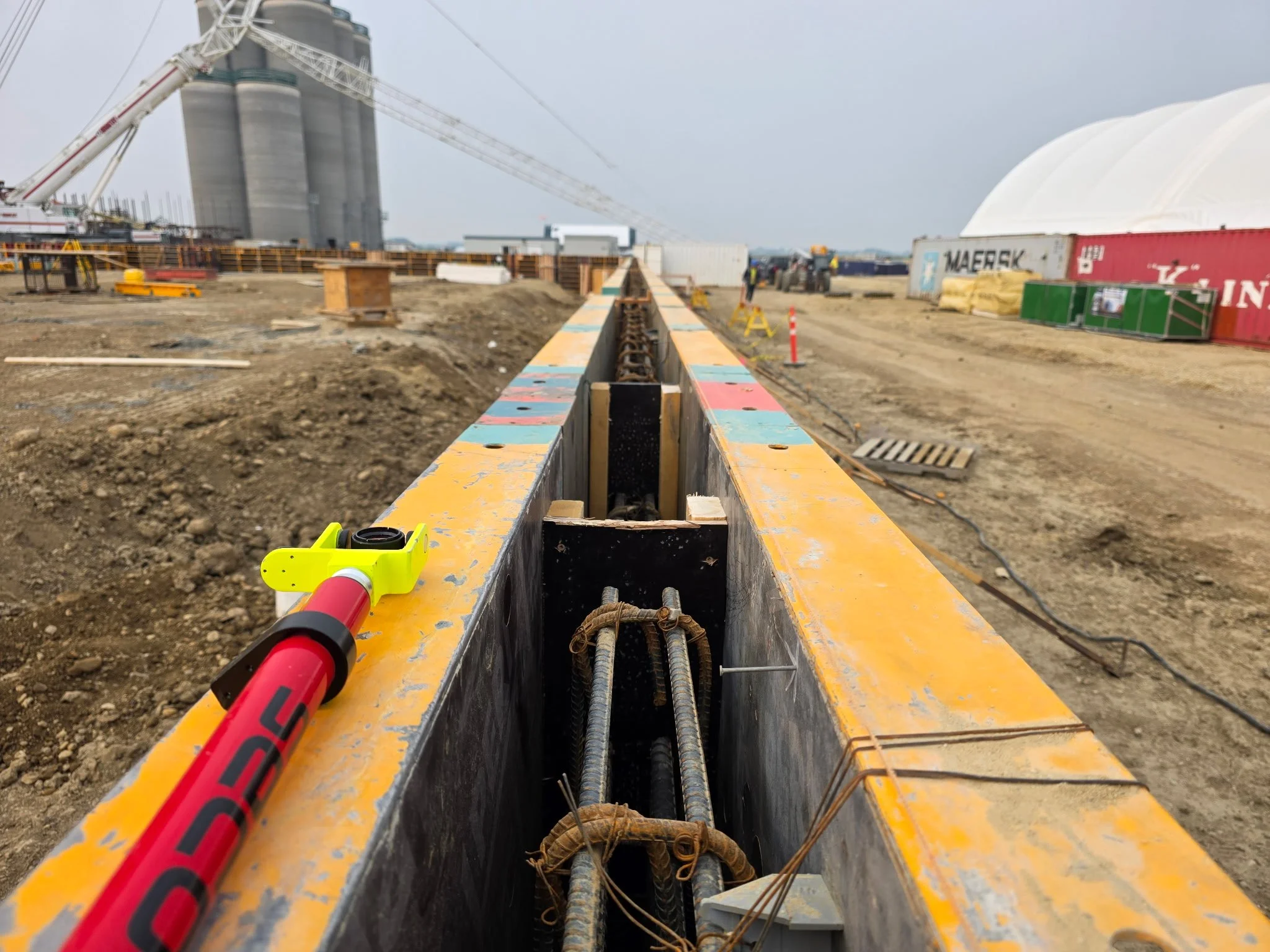 A construction site showing a railway track installation with a yellow metal frame, construction workers in the background, storage containers, and large industrial structures under a cloudy sky.